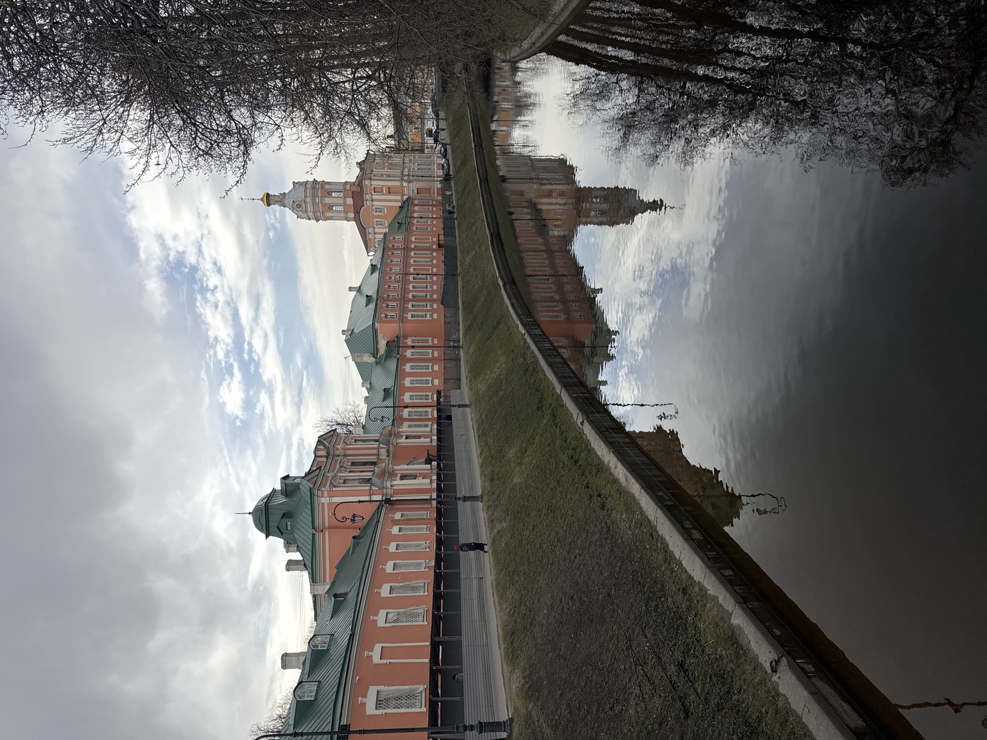 The image shows a view of the Alexander Nevsky Lavra monastery complex reflected in the Monastyrka River in Saint Petersburg, Russia. The buildings are light orange with green roofs and white window frames. A tall bell tower with a gold dome stands prominently in the center of the complex. A narrow strip of grass and a concrete embankment separate the buildings from the river. Bare trees line both the bank of the river, with their branches partially obscuring the view. The sky is overcast with light and dark gray clouds. A single person is visible walking on the pathway along the building.