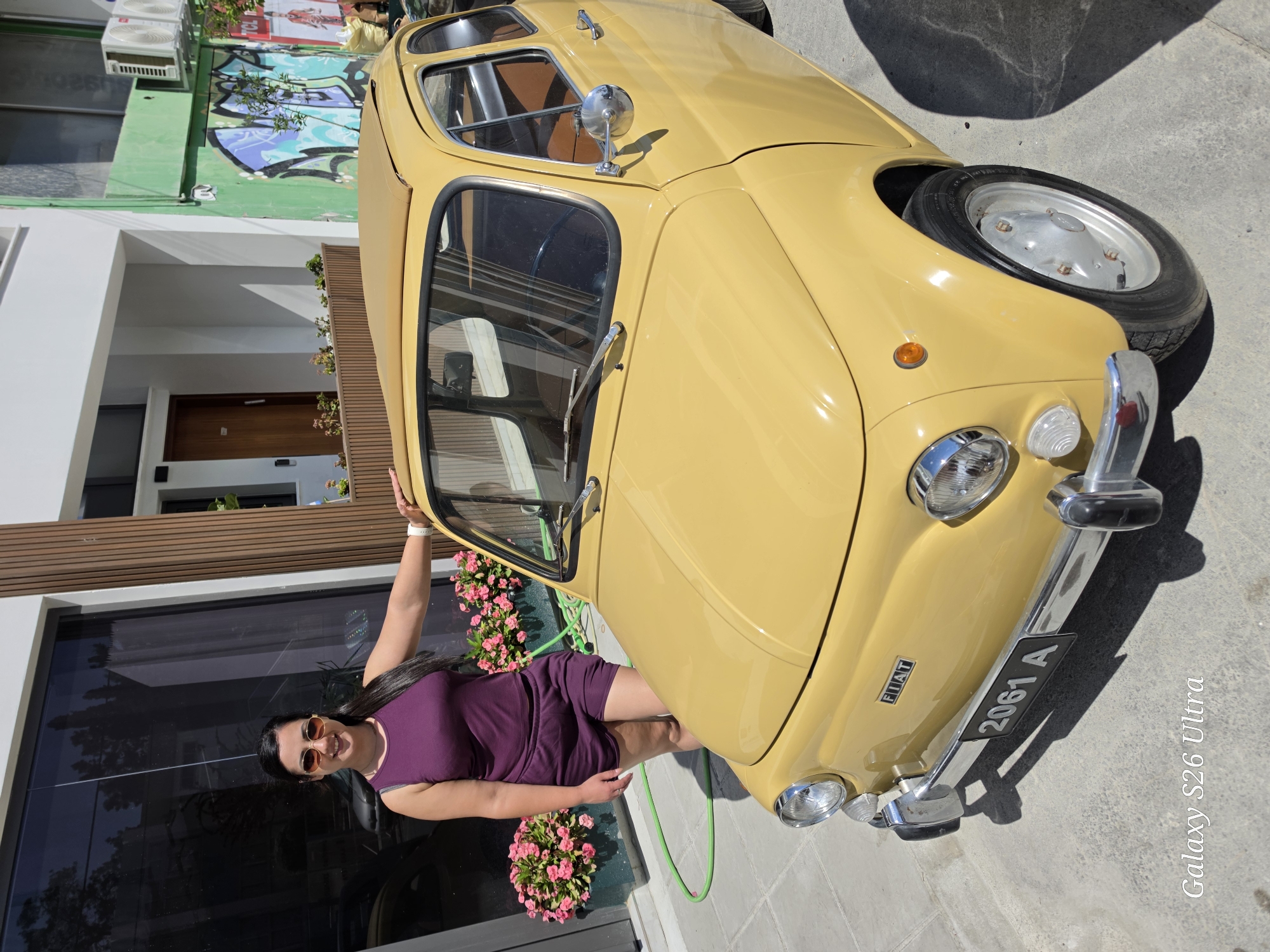 A woman stands beside a yellow Fiat 500 in Larnaca, Cyprus. She is wearing a short purple dress and sunglasses. The car has a license plate reading "2061 A" and a Fiat badge. A building with a brown door and flower boxes is in the background, along with graffiti on the upper part of the building. The ground is paved.