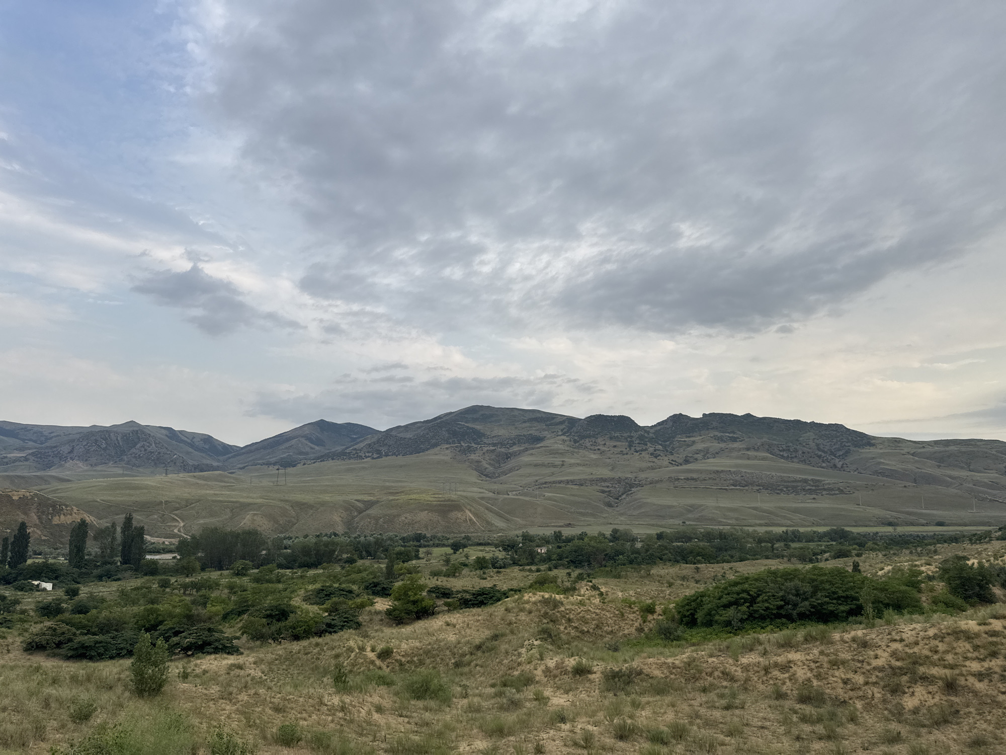 The image displays a panoramic view of a semi-arid landscape with rolling hills and a distant mountain range under a cloudy sky. In the foreground, there are sandy dunes covered with dry grasses and sparse vegetation. A middle ground features a dense cluster of green trees and shrubs, with a few taller, slender trees standing out. Beyond this, the landscape transitions into grassy hills with visible pathways and some scattered structures, possibly small buildings or power poles, extending towards the rugged, layered mountain peaks that dominate the horizon. The sky is overcast with varying shades of gray clouds, suggesting a subdued lighting condition. The scene is devoid of any human figures or explicit actions. The location is identified as being in the vicinity of Korkmaskala, Russia, implying a geographical context of a region with such topography.