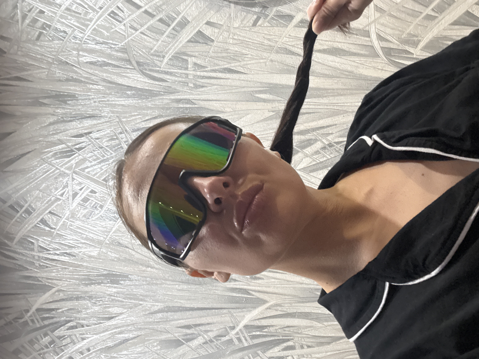 A medium close-up shot, originally rotated to the left, features a person, likely female, from the chest up in an indoor setting in Chaikovsky, Russia. The individual wears large, reflective sunglasses with a prominent rainbow tint across the lenses, obscuring their eyes. Their lips are slightly pursed or pouting. They are wearing a black top or shirt with distinct white piping along the neckline and possibly the sleeves, suggesting a pajama-like or casual style.
A hand is visible in the upper right corner, gently holding a dark, braided section of the person's hair, pulling it upwards. The background is a light-colored, possibly white or silver, textured wall covering, resembling a pattern of interwoven or feathery strands, creating a dynamic visual effect. The lighting is even, suggesting an indoor environment without strong natural light cues, making the time of day indeterminate. There is no visible text in the image. The overall impression is a casual, posed portrait.