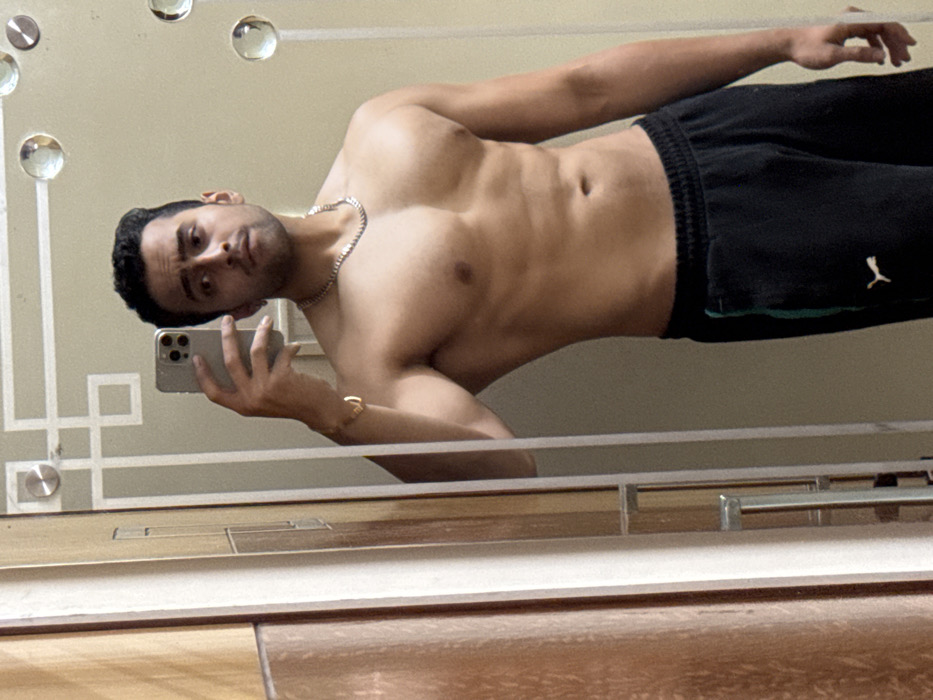 This image is a mirror selfie taken by a young man indoors, likely in Kolkata, India. The subject, who appears to be in his twenties or early thirties, is shirtless, revealing a muscular torso and abdomen. He has short dark hair, some facial stubble, and a neutral to slightly serious expression as he looks towards his reflection and the camera. He wears a silver chain around his neck and a gold ring on his left ring finger. His lower body is clad in black athletic pants or sweatpants, featuring a white Puma logo on the left thigh.
He holds a silver-colored smartphone, possibly an iPhone, in his left hand to capture the selfie. The mirror itself has decorative features, including multiple clear, spherical glass or plastic embellishments arranged in a pattern on the upper left side, and a geometric, squared-off design on the lower left. The background reflected in the mirror behind the man is a plain, light-colored wall. Below the mirror, the reflection shows what appears to be wooden flooring or a wooden surface, with a metallic bar or frame running horizontally across the bottom edge of the mirror's view. The lighting suggests an indoor setting.