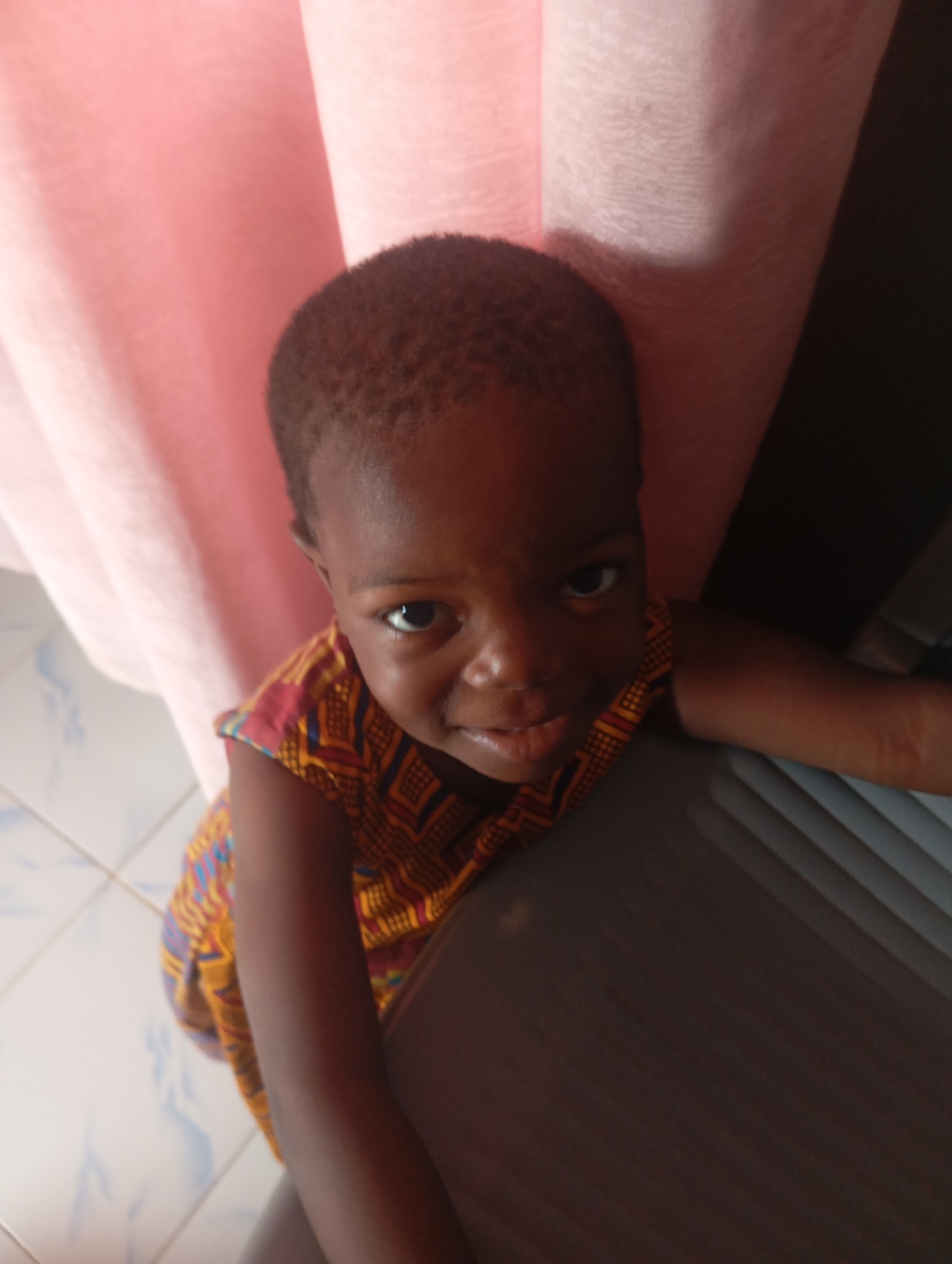 An upward-angle, eye-level medium shot captures a young child with dark skin, short hair, and dark eyes looking directly at the camera with a slightly concerned expression. The child is wearing a colorful, sleeveless garment with a geometric pattern in shades of yellow, orange, red, and blue. They are leaning against a dark gray object, possibly a piece of furniture or luggage, which occupies the right side of the frame. Behind the child, a large, soft, pink fabric drapes down from the top of the image, creating a blurred background. To the left of the child, a tiled floor with a light blue and white pattern is visible. The scene appears to be indoors.
