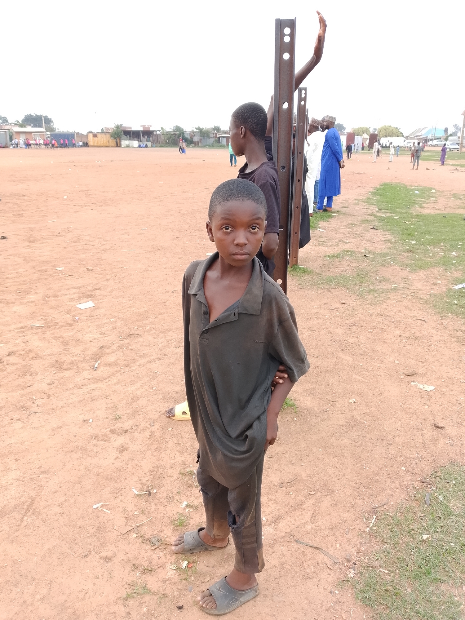 A young boy with dark skin and short hair stands in the foreground, looking directly at the camera. He is wearing a dark, oversized polo shirt and ripped dark pants, with gray sandals on his feet. He stands on a dusty, reddish-brown ground with scattered debris and sparse patches of dry grass. Behind him, a metal pole stands vertically, and further back, another young person is partially visible, with their arm raised. In the background, several adults are gathered, some wearing traditional clothing, including a blue robe and a white garment. There are buildings, fences, and vehicles in the distance, suggesting an outdoor event or gathering in an urban or semi-urban environment. The sky is overcast and white.