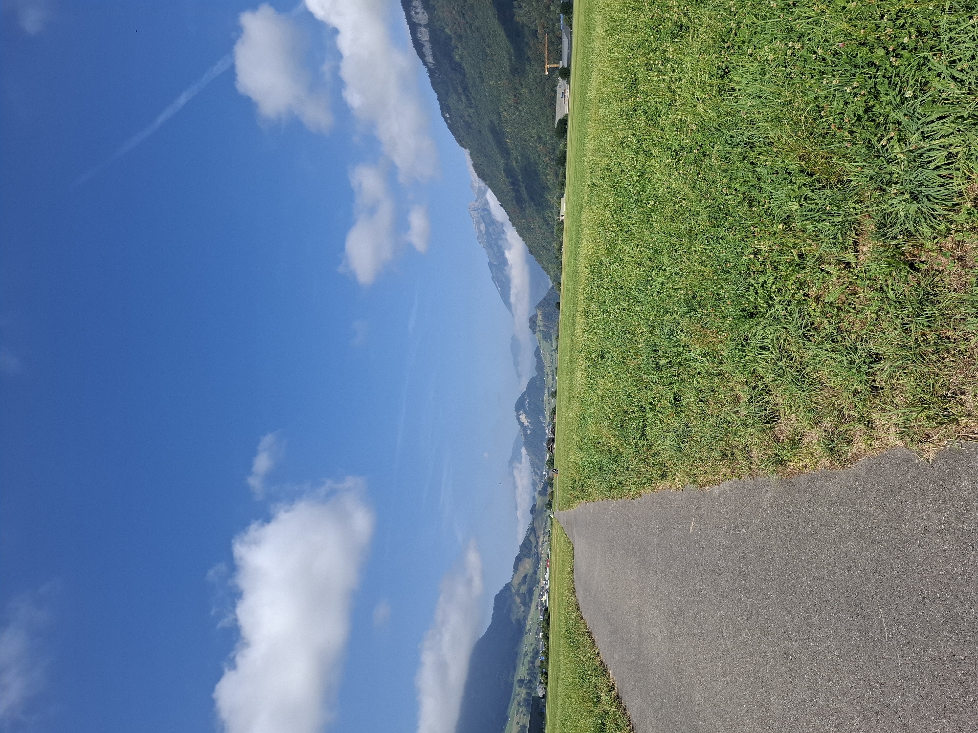 A paved path curves through a vibrant green field under a bright blue sky dotted with fluffy white clouds. The path leads towards a distant landscape of rolling hills and majestic mountains. In the distance, buildings and a construction crane are visible, suggesting a rural or semi-rural setting. The overall scene captures the serene beauty of the Swiss countryside, likely in the vicinity of Buochs, Switzerland, given the characteristic alpine landscape.
