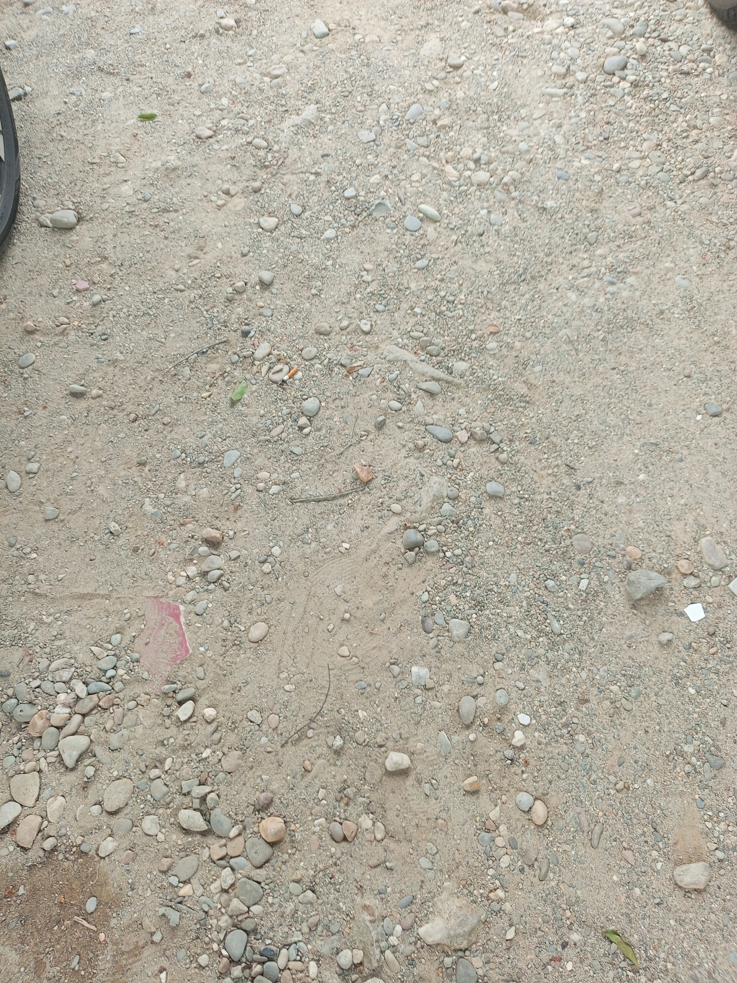 The image depicts a close-up view of a gravelly, sandy ground surface. The ground is covered with a variety of small to medium-sized pebbles and stones, interspersed with loose sand and dirt. A fragment of pink material, possibly paper or plastic, is visible on the ground in the lower left quadrant of the image. There are also a few scattered pieces of debris, including what appears to be a cigarette butt with an orange filter in the upper right quadrant, and a single green leaf towards the center left. A portion of a dark tire tread is visible on the far left edge of the image, suggesting it may be from a vehicle. The overall impression is of an outdoor, unpaved area. There is no indication of specific landmarks or features that would pinpoint the location within the city of Langsa, Indonesia.