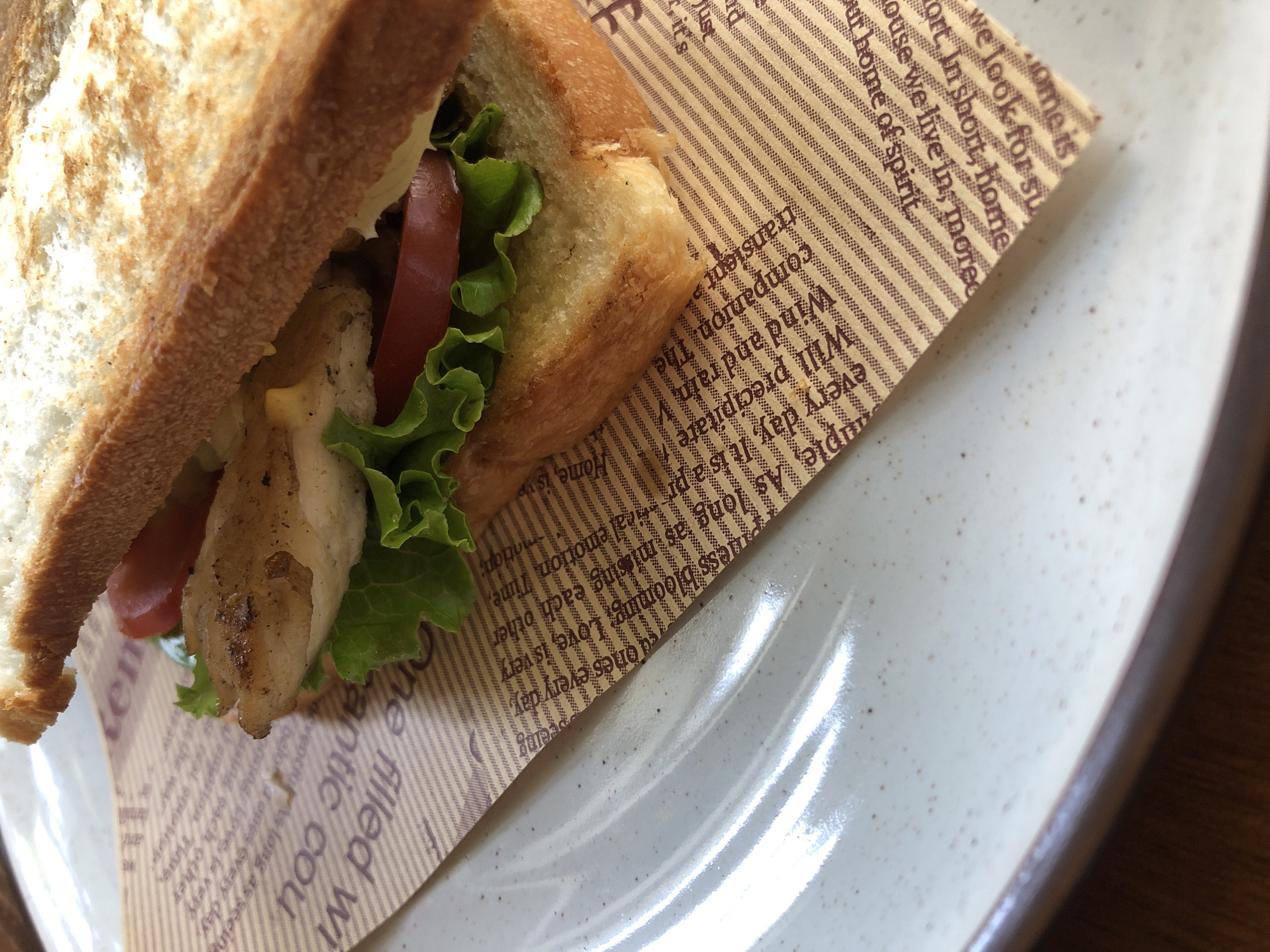 A close-up, low-angle shot shows a sandwich on a plate in Da Nang, Vietnam. The sandwich is made with toasted bread, grilled chicken, lettuce, and tomato. It is served on a piece of brown paper with a newspaper print. The plate is white and has a speckled pattern. The background is blurred, but appears to be a wooden table. The lighting is soft and natural, creating a warm and inviting atmosphere. The scene is very clean and organized.