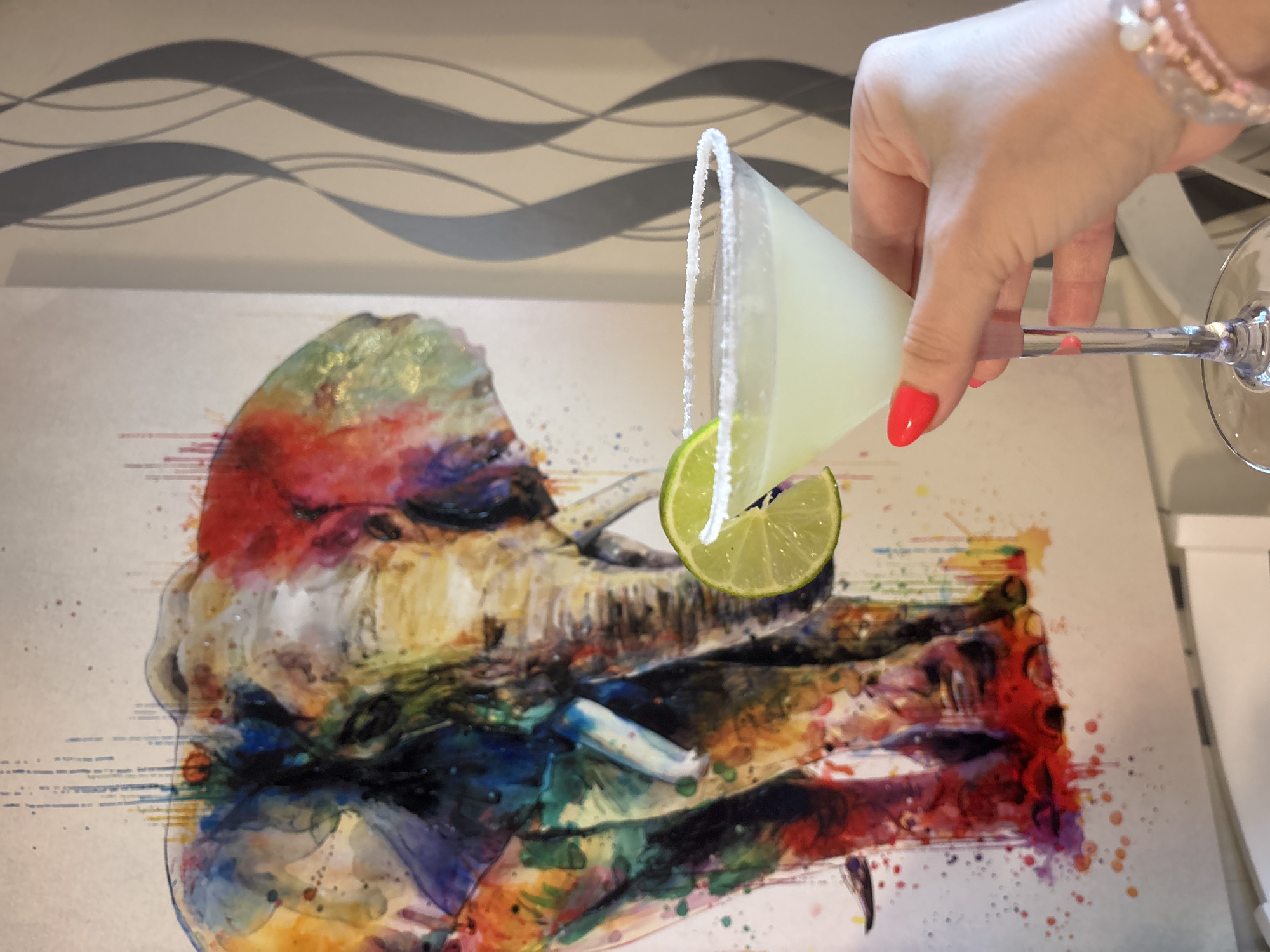 A photograph taken indoors, likely in a domestic setting, shows a person's hand holding a margarita glass with a salted rim and a slice of lime on the edge. The glass contains a pale yellow liquid. The person has red nail polish and is wearing a bracelet.
Below the glass, there's an abstract watercolor painting of a person; red, blue, and yellow are the dominant colors. The background features a white surface with light grey wavy lines. A portion of a white cabinet is visible in the upper-right corner of the photo.
There is no information within the image to establish the location within the city of Sofia, Bulgaria.