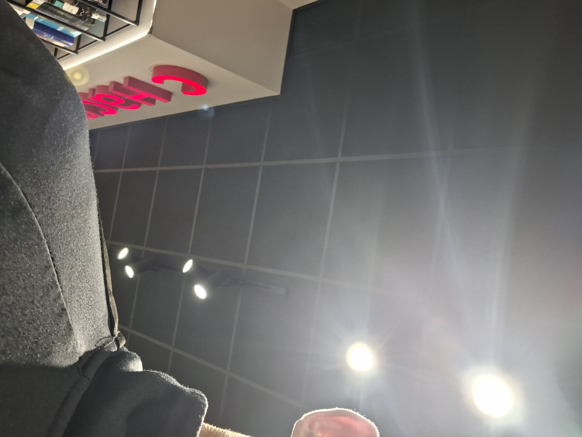 The image depicts an indoor commercial environment, observed from a low-angle perspective looking upwards. In the lower right quadrant, the upper portion of a human shoulder, clad in a dark, possibly fleece-textured garment, is visible. To the immediate right, the top of a human ear, brightly backlit and appearing somewhat translucent, is partially captured.
The ceiling consists of dark grey square grid tiles, characteristic of a commercial suspended ceiling. Multiple circular recessed downlights are prominent, emitting intense artificial illumination that generates significant lens flares and light streaks across the upper and central portions of the frame. Towards the mid-left, a light-colored, possibly off-white, architectural surface is visible, upon which large, three-dimensional letters in a vibrant pink or red hue are mounted. The visible letters clearly spell "Cho," indicating part of a larger sign, likely "Chocoland" or a similar confectionery brand. Below and to the left of this signage, a segment of a dark metal display shelf holding various bottles or containers can be seen.
Based on the distinctive "Cho" signage, the location is identified as a Chocoland store within a shopping mall in Varna, Bulgaria. The overall scene suggests a retail setting with modern lighting and commercial display elements.
