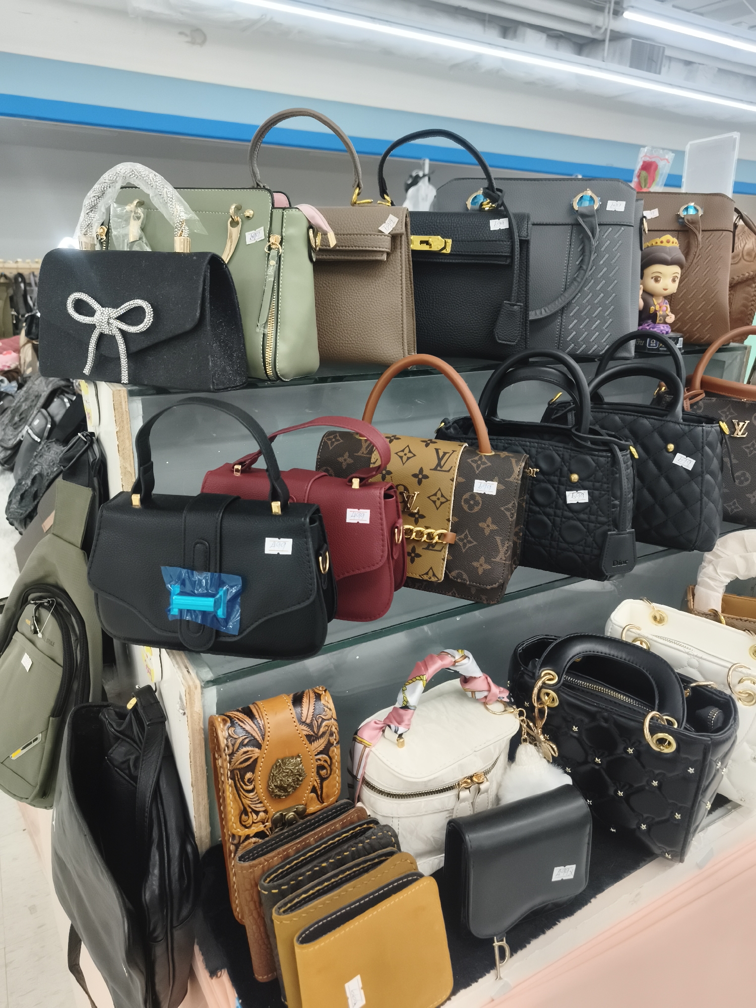 The image shows a display of handbags inside a store, likely in บ้านบางรักษ์, Thailand. The handbags are arranged on shelves. There's a variety of styles and colors, including black, brown, olive green, and red. Some bags feature logos or embellishments like bows, studs, and quilted patterns. Wallets are also visible on the lower shelves, arranged next to the bags. The background indicates a typical retail environment with shelves and overhead lighting. A small doll is placed between two handbags.