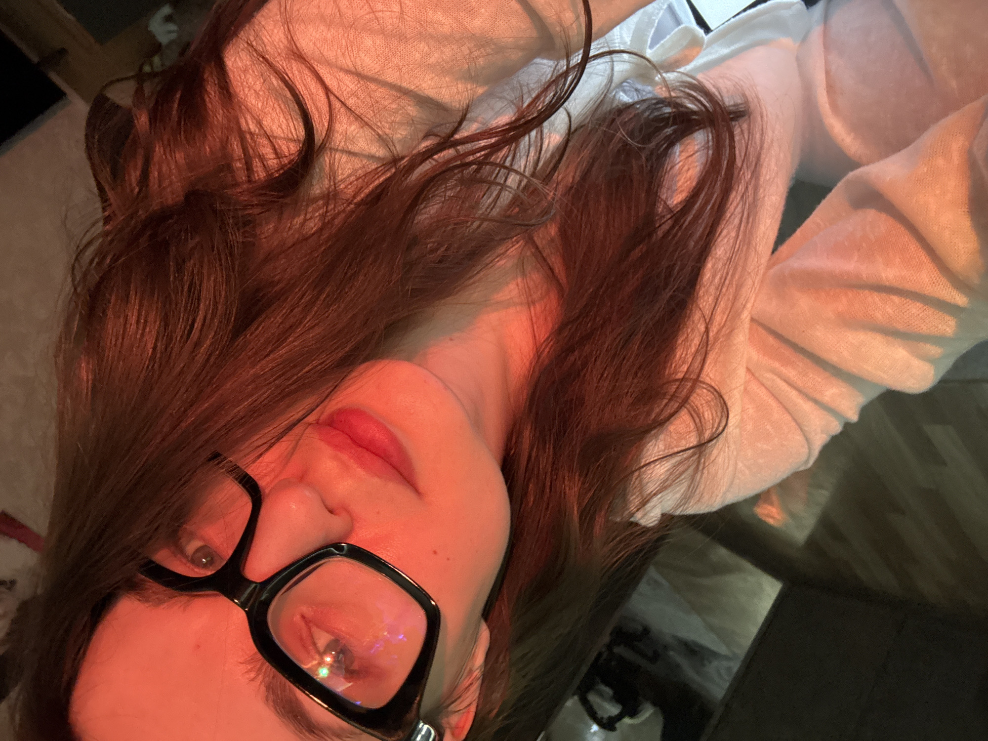 A selfie of a woman with long brown hair and glasses. She is wearing a light pink or white sweater. The image is lit with a reddish or orange hue. The background is slightly blurred and shows a dark wooden floor and glimpses of other indistinct objects. There is no evidence to indicate this image was taken in the city of Chaikovsky, Russia.