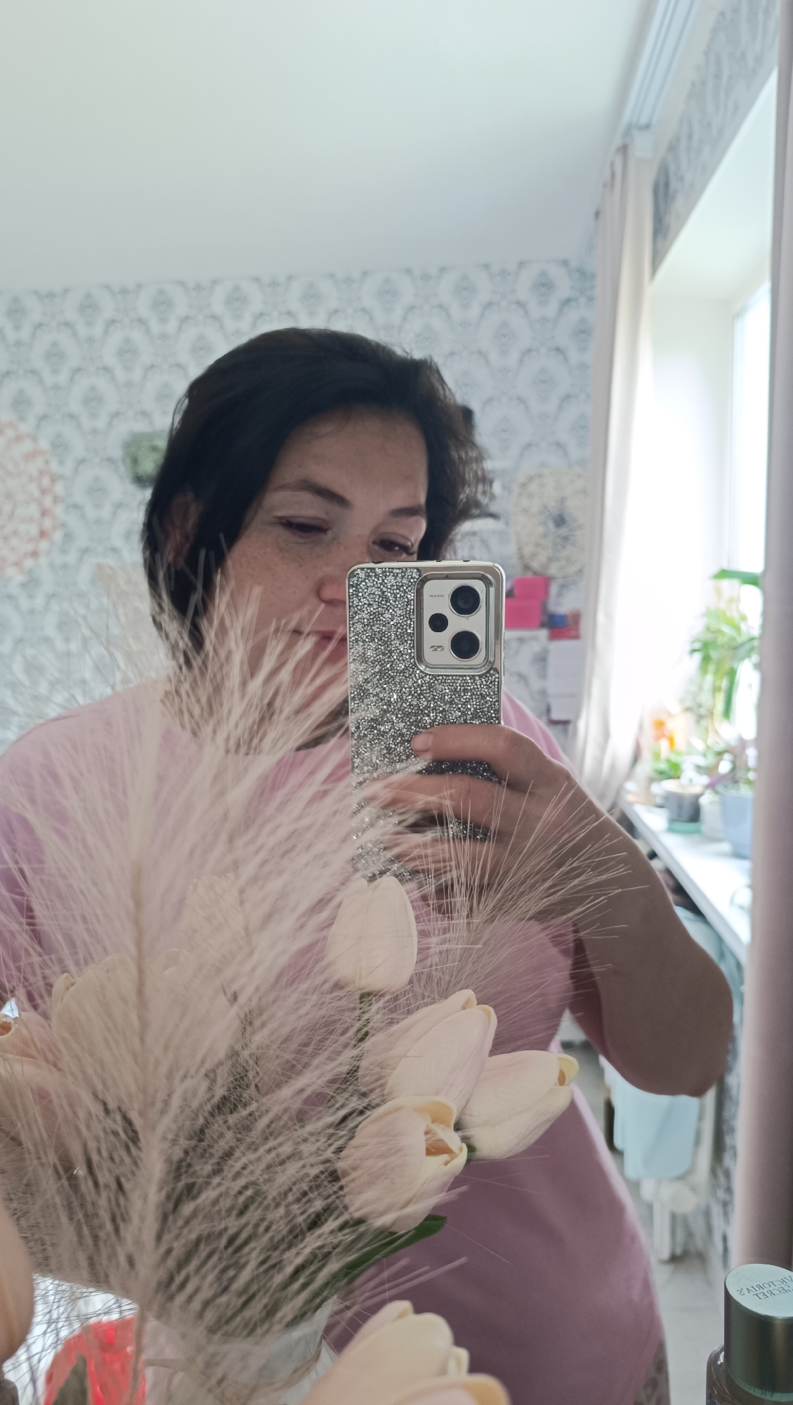 A woman with dark hair and freckles is taking a mirror selfie in what appears to be a residential setting, possibly an apartment in Chaikovsky, Russia. She is wearing a pink t-shirt and holding a bouquet of pale pink tulips and fluffy white feathers in front of her. Her phone, with a sparkly silver case, is visible in her hand as she takes the picture. The background shows patterned wallpaper, a window with light-colored curtains, and a shelf with plants and decorative items.