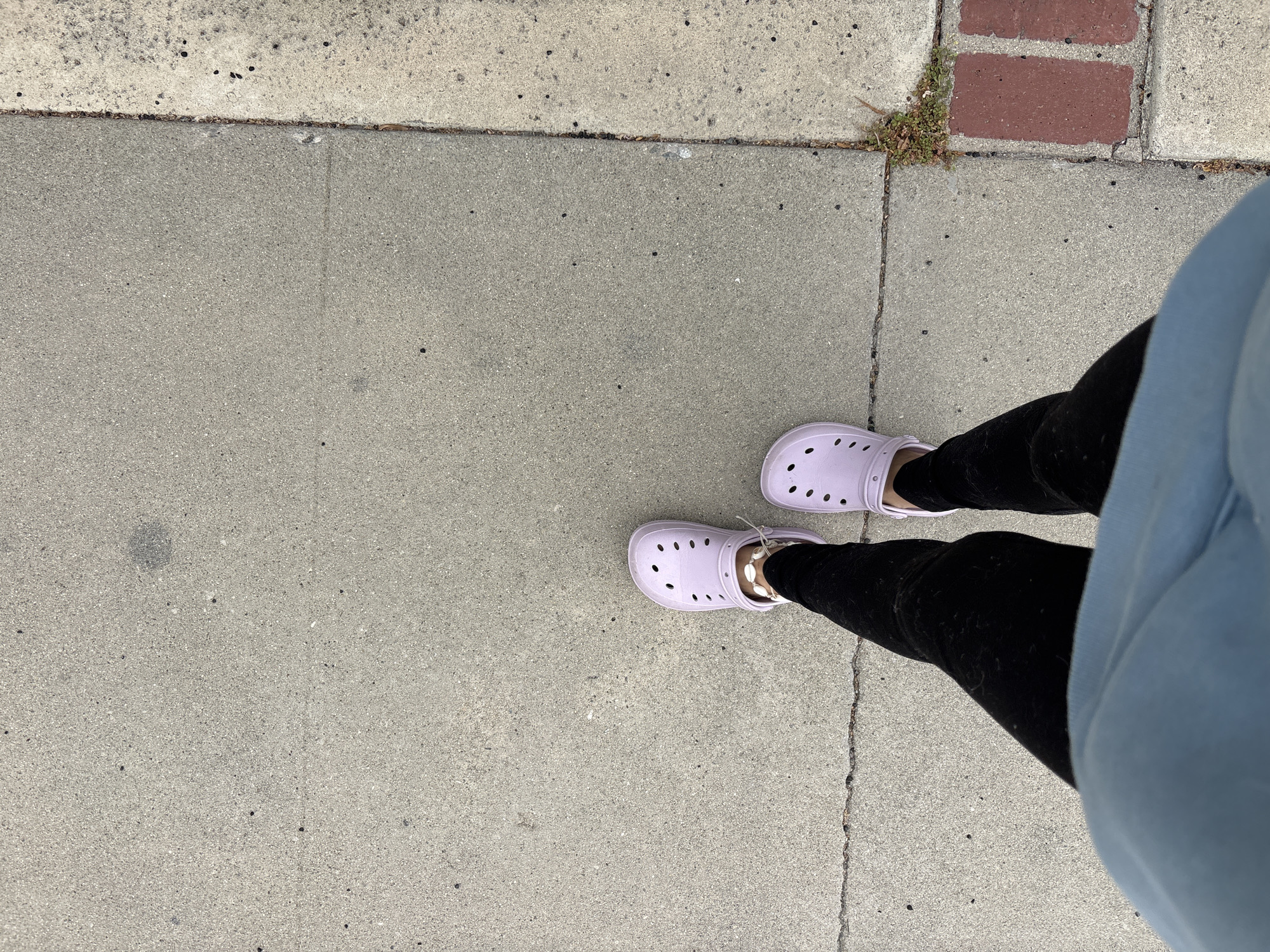 The image shows a first-person perspective of someone standing on a concrete sidewalk in Seal Beach, California. The person is wearing black leggings and lavender Crocs. A blue sweatshirt partially obscures the right side of the frame. The sidewalk is cracked in places and abuts a different colored section of concrete and a small area with red brick. There are some small weeds growing in the cracks near the brick area.