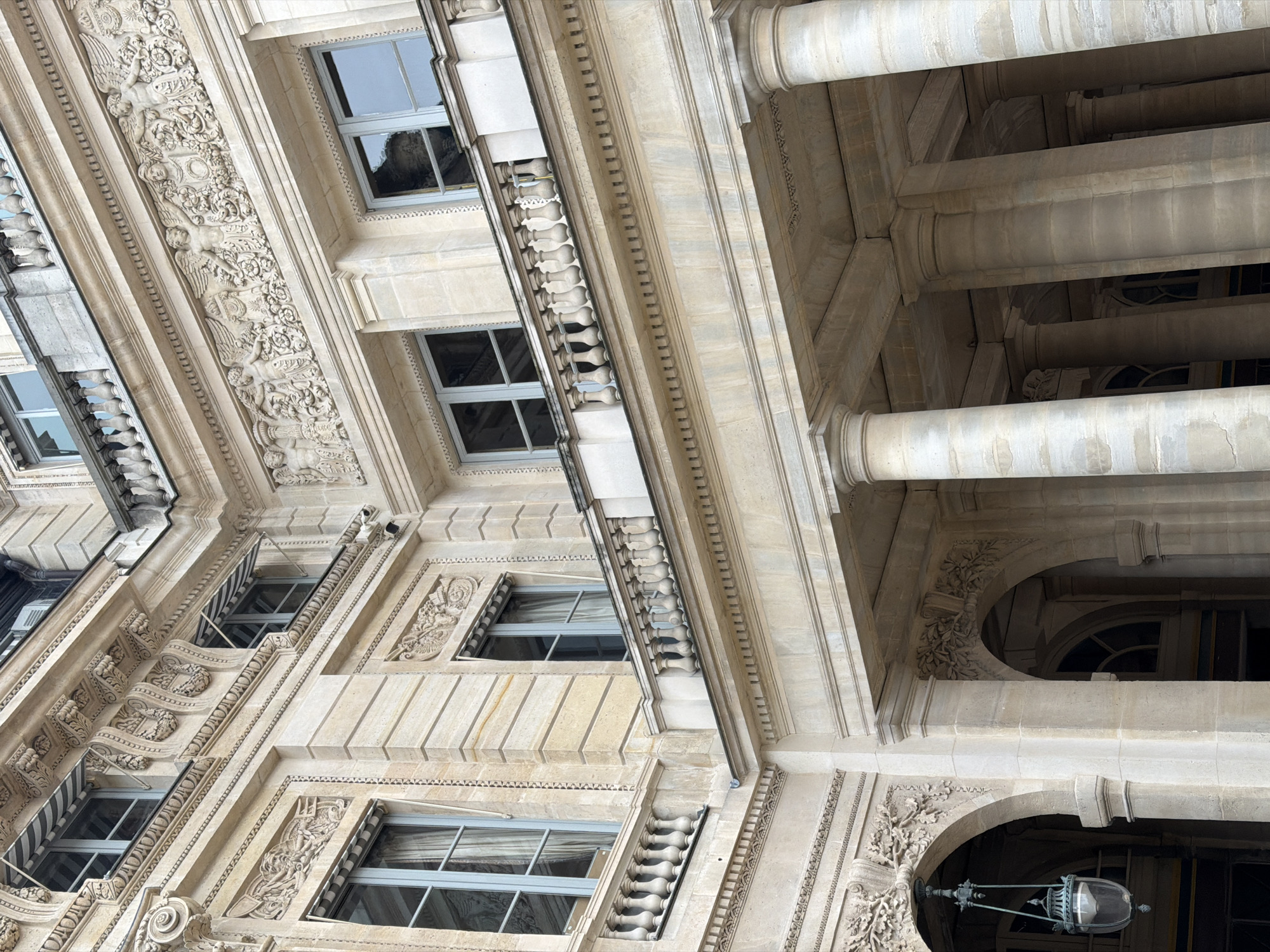 The image depicts the exterior architecture of a building, likely located in Paris, France. The building features classical elements such as columns, ornate carvings, and windows with detailed frames. The material of the building appears to be a light-colored stone, such as limestone or marble. The windows suggest multiple stories, with some having awnings. There is an arched entryway at the bottom right corner, adorned with floral carvings and a vintage-style lamppost hanging nearby. The perspective is from below, looking upward at an angle, emphasizing the scale and grandeur of the building's facade.