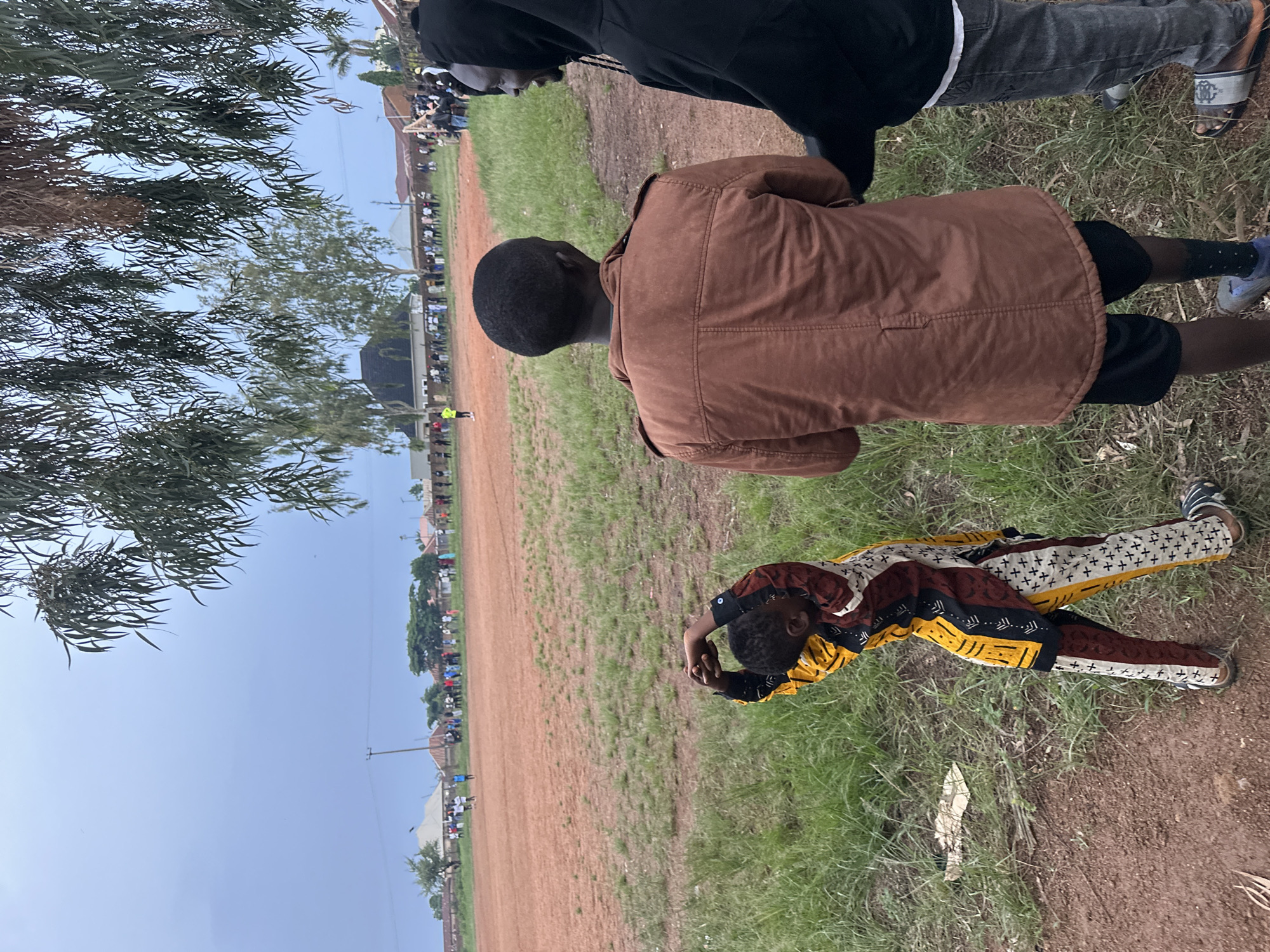 A group of people are gathered on a dirt field in Du, Nigeria, under an overcast sky. In the foreground, a young child wearing a colorful patterned outfit stretches their arms above their head. To the right, two men are standing, facing away from the camera. The man in the middle wears a brown jacket, while the man to his right is in a black hoodie. Beyond them, a line of people stands at the edge of the field, and a few individuals can be seen on the field itself. Trees and buildings are visible in the background, suggesting a semi-urban environment. The ground is a mixture of dirt and sparse green grass.