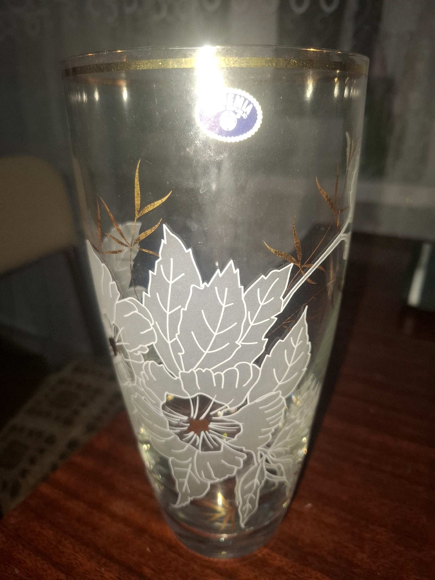 A clear glass vase with a gold rim and a white floral design is centered in the frame, resting on a dark wooden surface. The vase features an etched pattern of large, stylized white leaves and smaller, delicate gold branches with leaves. A circular blue and white sticker with text is visible on the upper portion of the vase, partially obscured by a bright light reflection. The background is dark and out of focus, with hints of furniture and possibly a window. The lighting suggests an indoor setting, likely at night, with a strong light source creating glare on the glass. There are no people or animals present. This image was taken in Almaty, Kazakhstan.