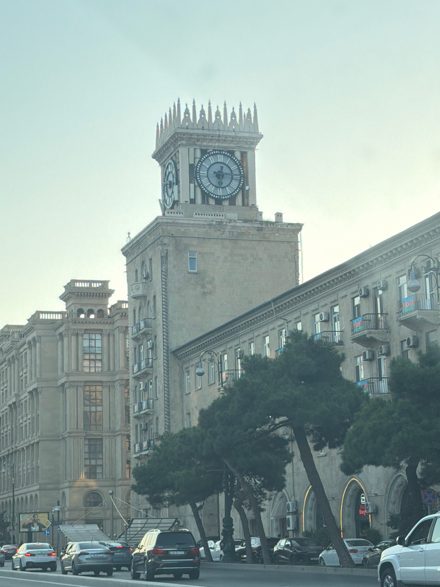 The image shows a street scene in Baku, Azerbaijan, with a prominent clock tower building. The clock tower has a decorative crown and two large circular clock faces. Several multi-story buildings with architectural details like balconies, columns, and arched windows line the street. Some of the buildings display the Azerbaijani flag on their balconies. Trees are present along the sidewalk, some with leaning trunks. Vehicles, including cars and an SUV, are driving on the road. The sky is overcast and a pale blue. The scene appears to be a daytime shot of a commercial or civic area in the city.