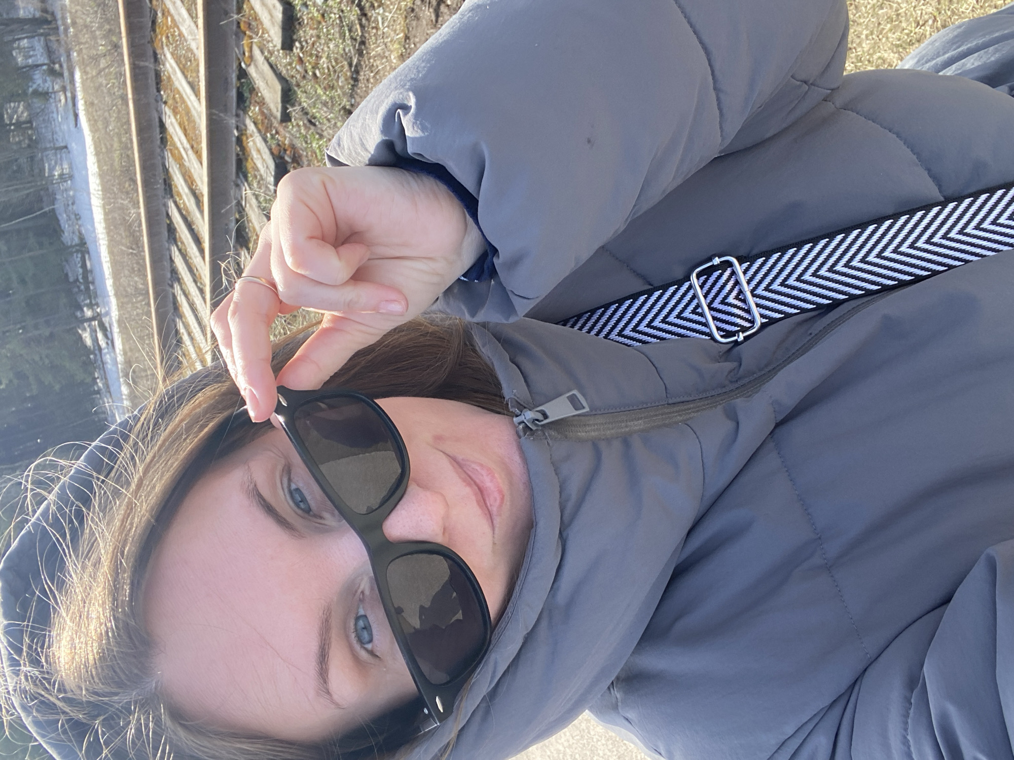 A woman in a gray puffer jacket with a black and white chevron strap is seen holding sunglasses, likely taking a selfie outdoors. In the background, there are train tracks. The location is Chaikovsky, Russia.