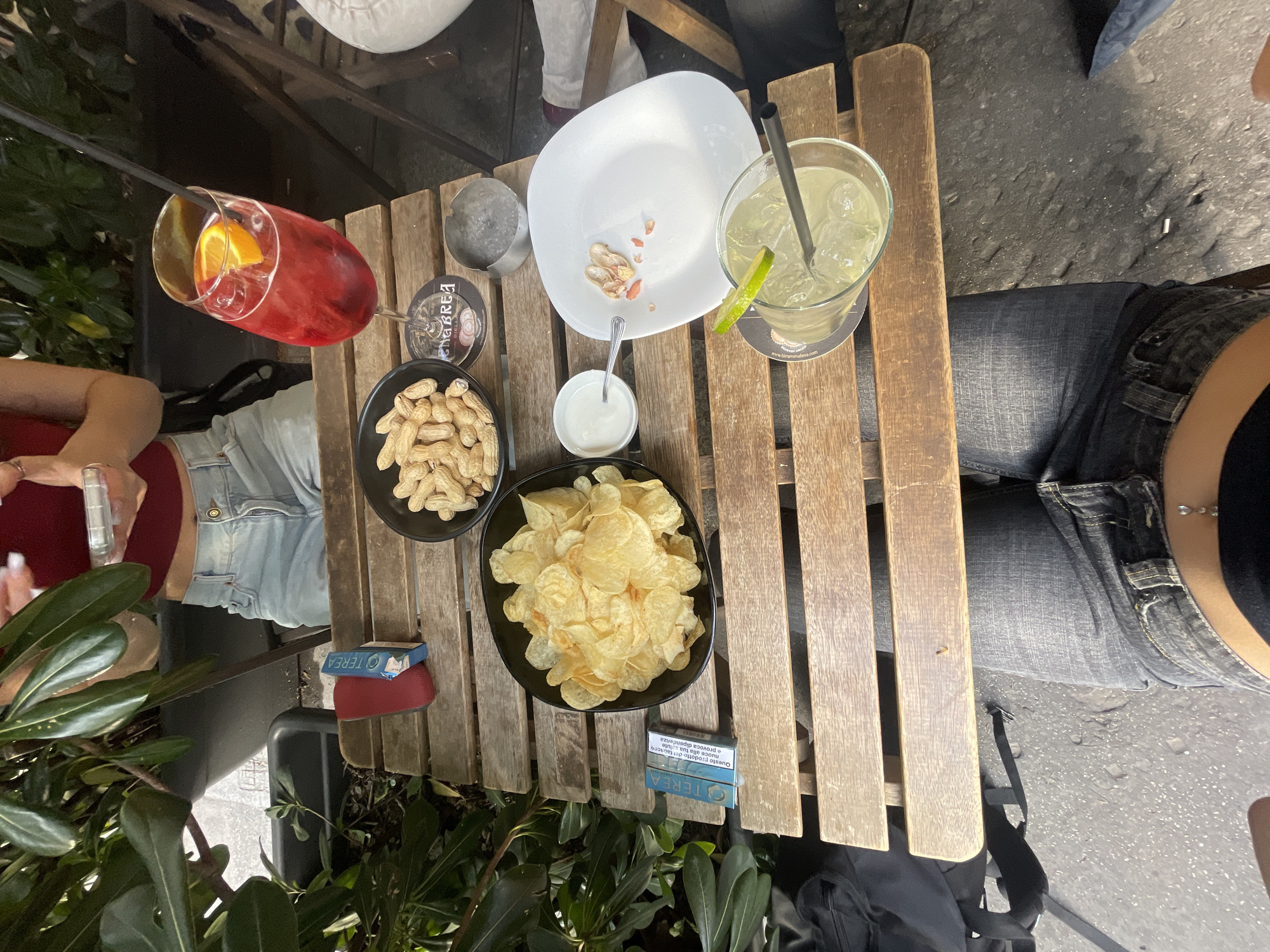 Here is a forensic description of the image:
**Scene:** An outdoor table setting, likely at a cafe or bar. The table is made of wood slats with gaps between them. The overall impression is casual and informal.
**Location:** The presence of "TEREA" brand cigarettes and "CHABREA BIELLA" on a drink coaster suggests the location is in Italy, possibly the city of Milan.
**People:** Two or three people are partially visible. One person is wearing a denim skirt with a red top and holding a vaping device in their left hand. Another is wearing gray jeans.
**Objects:**
* Aperol Spritz (red cocktail with orange slice)
* A clear glass of a light-colored drink with ice and a lime garnish, likely a Gin and Tonic.
* A white square plate containing some discarded peanut shells
* A small metal ice bucket.
* A small bowl of peanuts
* A small bowl of white dip
* A large bowl of potato chips
* A pack of cigarettes.
* A red card holder.
* A black backpack
**Actions and Interactions:** People are engaging in a social activity, likely drinking and eating at the table.
**Environment:** There are plants present, some partially out of focus, suggesting the table is near a garden or landscaping. The ground is paved.