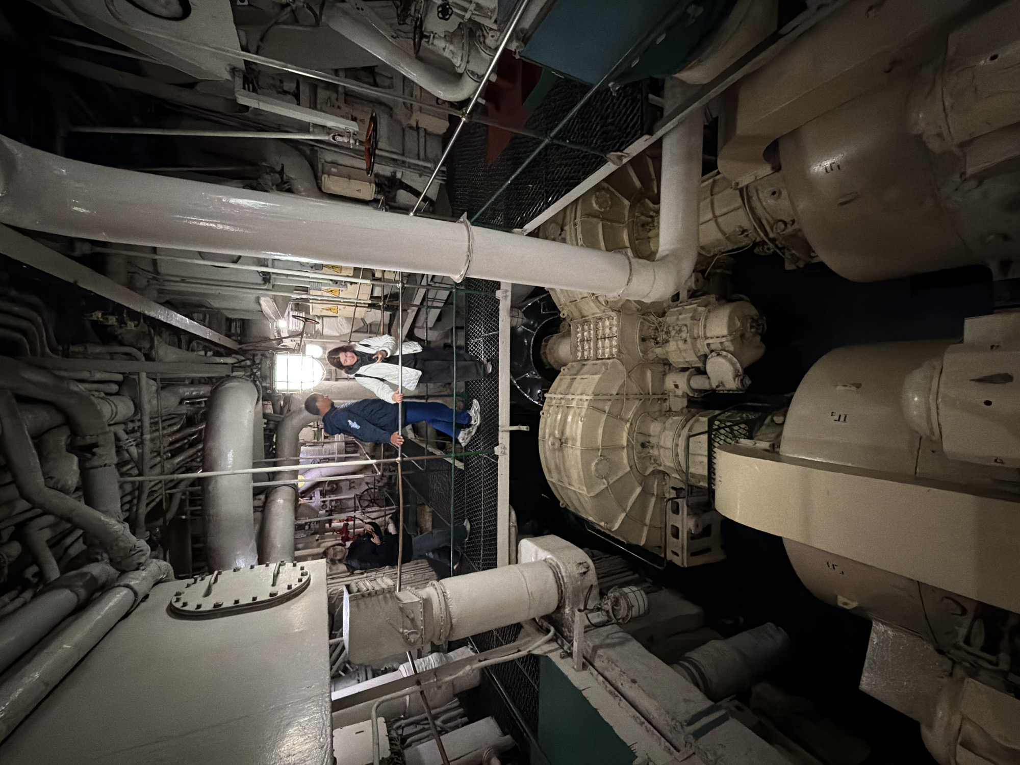 In Murmansk, Russia, inside the engine room of the nuclear icebreaker *Lenin*, three individuals are observed standing on a metal walkway overlooking a massive piece of machinery. The walkway is a dark gray, grated metal structure with a green handrail. The engine room is characterized by its industrial complexity, filled with a dense network of pipes, conduits, and large metallic components. To the left, a person in a black jacket and jeans stands looking at their phone, with their back to the camera. To their right, a man in a blue jacket and jeans is looking down into the engine room. Further to the right, a woman in a white jacket and dark pants is also looking down, holding a phone in her hand. The primary focus of the lower part of the image is a large, beige, turbine-like machine with visible components and branding. The environment is dimly lit, with industrial lighting fixtures casting a stark illumination on the machinery and walkways.