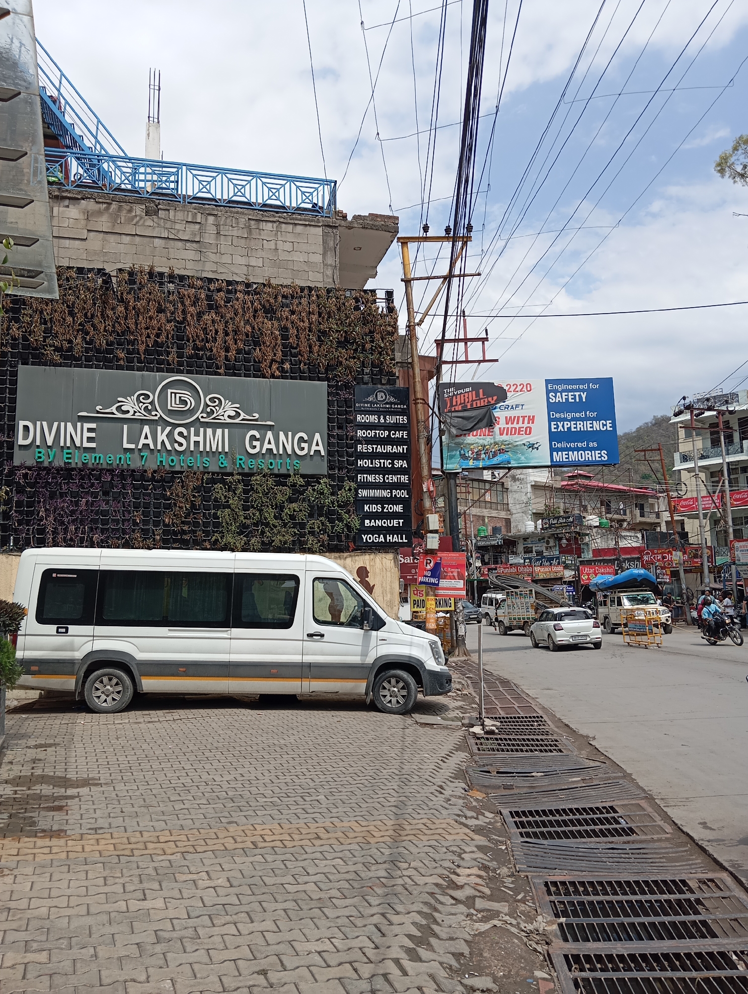 The image depicts a street scene in Jonk, India. In the foreground, a white passenger van with a yellow stripe is parked on a brick-paved area beside a street with metal drain grates. Behind the van is a building with a green wall covered in vegetation and a large sign that reads "DIVINE LAKSHMI GANGA By Element 7 Hotels & Resorts," listing amenities. The street is busy with vehicles including cars, auto-rickshaws, and motorcycles, as well as some pedestrians. Billboards advertise the Shivpuri Thrill Factory and safety-engineered experiences. Various shops and businesses line the street, with signs in both English and Hindi, including "Punjabi Dhaba," and "Uttar Dakshin." Power lines and utility poles are visible overhead.