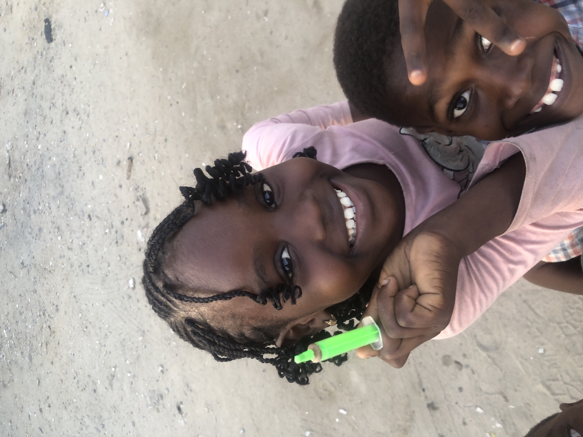Two young Black children, a girl and a boy, are positioned in the foreground of the image, looking upwards at the camera. The girl, on the left, has braided hair and is wearing a pink t-shirt. She is holding a small green object, possibly a toy or a novelty item, in her right hand. She is smiling broadly, with her teeth visible. The boy, on the right, has short, dark hair and is wearing a plaid shirt. He is also smiling and appears to be making a gesture with his right hand, with two fingers extended towards the camera. The background is a sandy, textured surface, suggesting an outdoor environment. This scene is likely located in Ado, Nigeria.