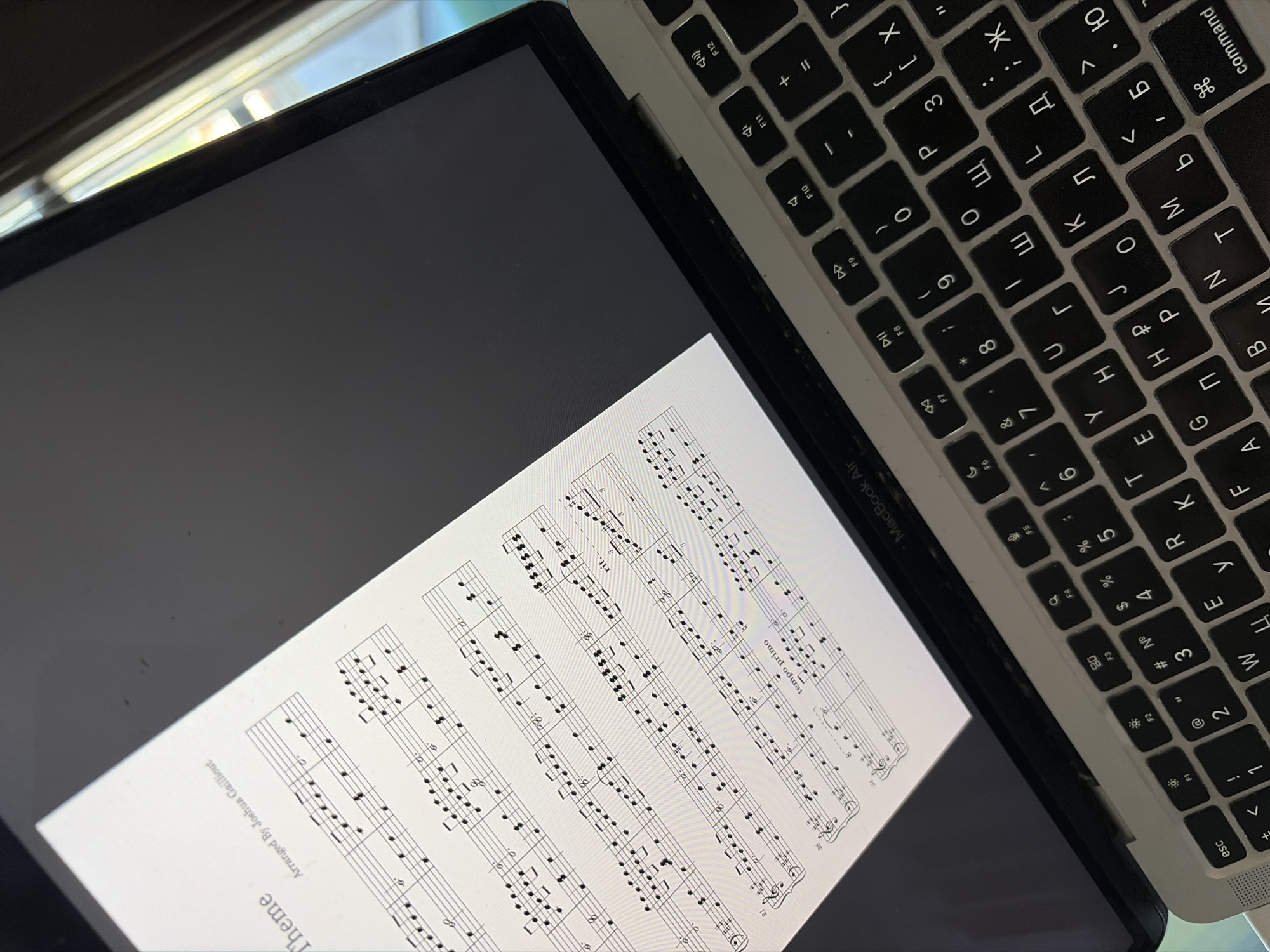 The image shows a laptop displaying sheet music on its screen. A portion of the keyboard is visible, featuring both English and Cyrillic characters. The sheet music title is "Theme," arranged by Joshua Gailliout. A partial label on the laptop identifies it as a "MacBook Air." The environment appears to be indoors, with daylight present from an unseen window. There is no indication of this image being located in Budva, Montenegro.