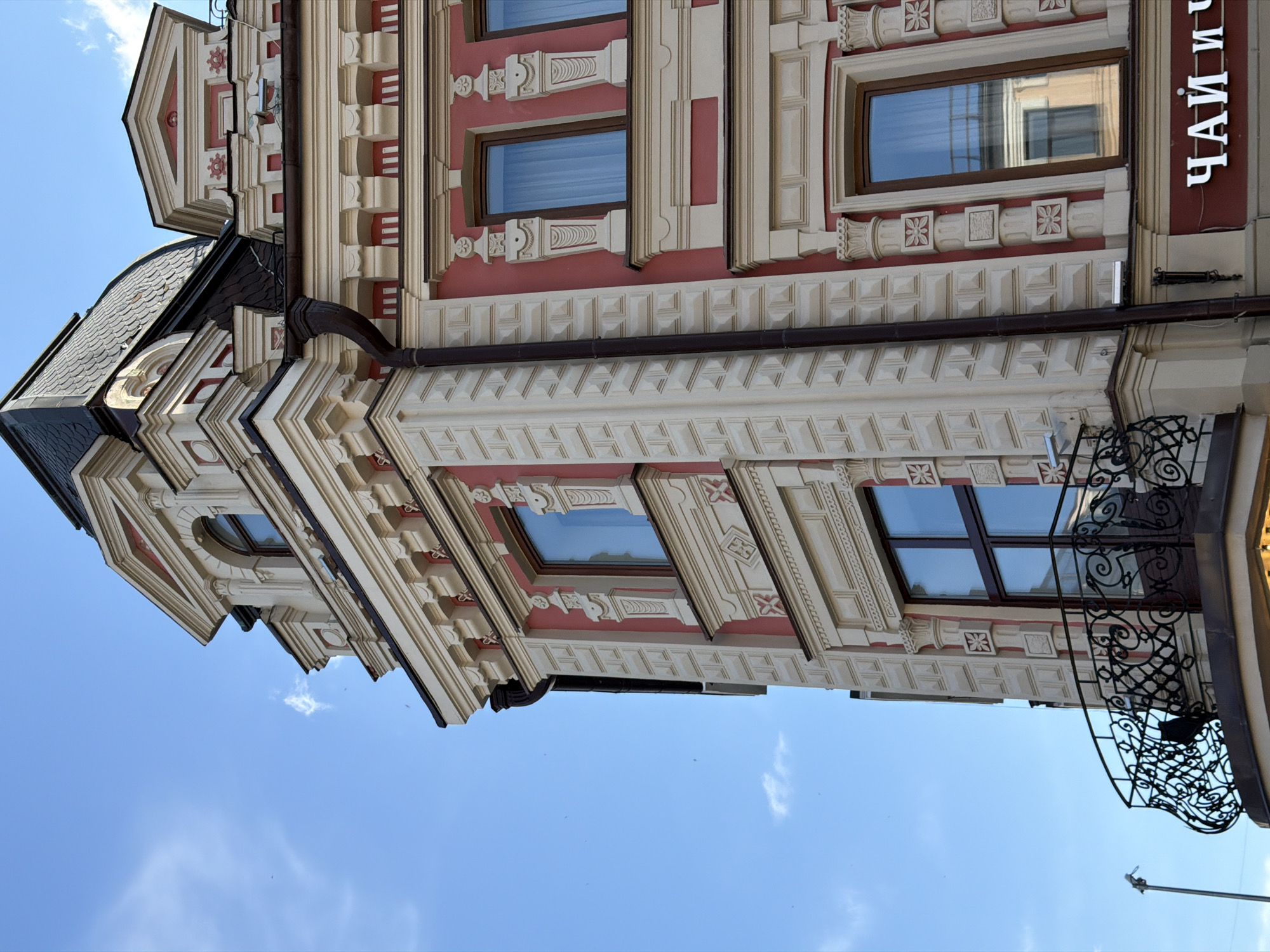 An upward-angled view captures the ornate corner of a multi-story building against a bright blue sky with wispy white clouds. The building's main wall surfaces are a salmon-pink/reddish hue, contrasted by extensive creamy/light beige architectural details.
The prominent corner features stacked, rectangular quoins in the lighter beige. Above, a dark, mansard-style roof with scale-like tiles includes a dormer window topped by a pediment and an arched window. Another pediment is visible further along the roofline to the right. Several dark brown drainpipes are affixed vertically along the corner.
Multiple rectangular windows with dark brown frames are visible; their glass reflects the sky and elements of an opposing structure. The windows are flanked by decorative pilasters and feature ornate sills and cornices. Various decorative panels are interspersed, showcasing floral, geometric (e.g., 'X' shapes), and classical motifs.
A curved, black wrought-iron balcony with intricate scrollwork is positioned on a lower floor, wrapping around the building's corner. No individuals are present on the balcony. On the building's reddish wall, near the bottom right, white Cyrillic letters are partially visible, spelling "ЧАЙ И Ч", indicating a commercial establishment likely related to tea. A slender, light-colored pole extends into the sky on the far left. Small, dark specks, likely birds or insects, are visible against the sky. The scene is illuminated by bright daylight.
This architectural style and Cyrillic signage are consistent with a location in Kazan, Russia, as specified.