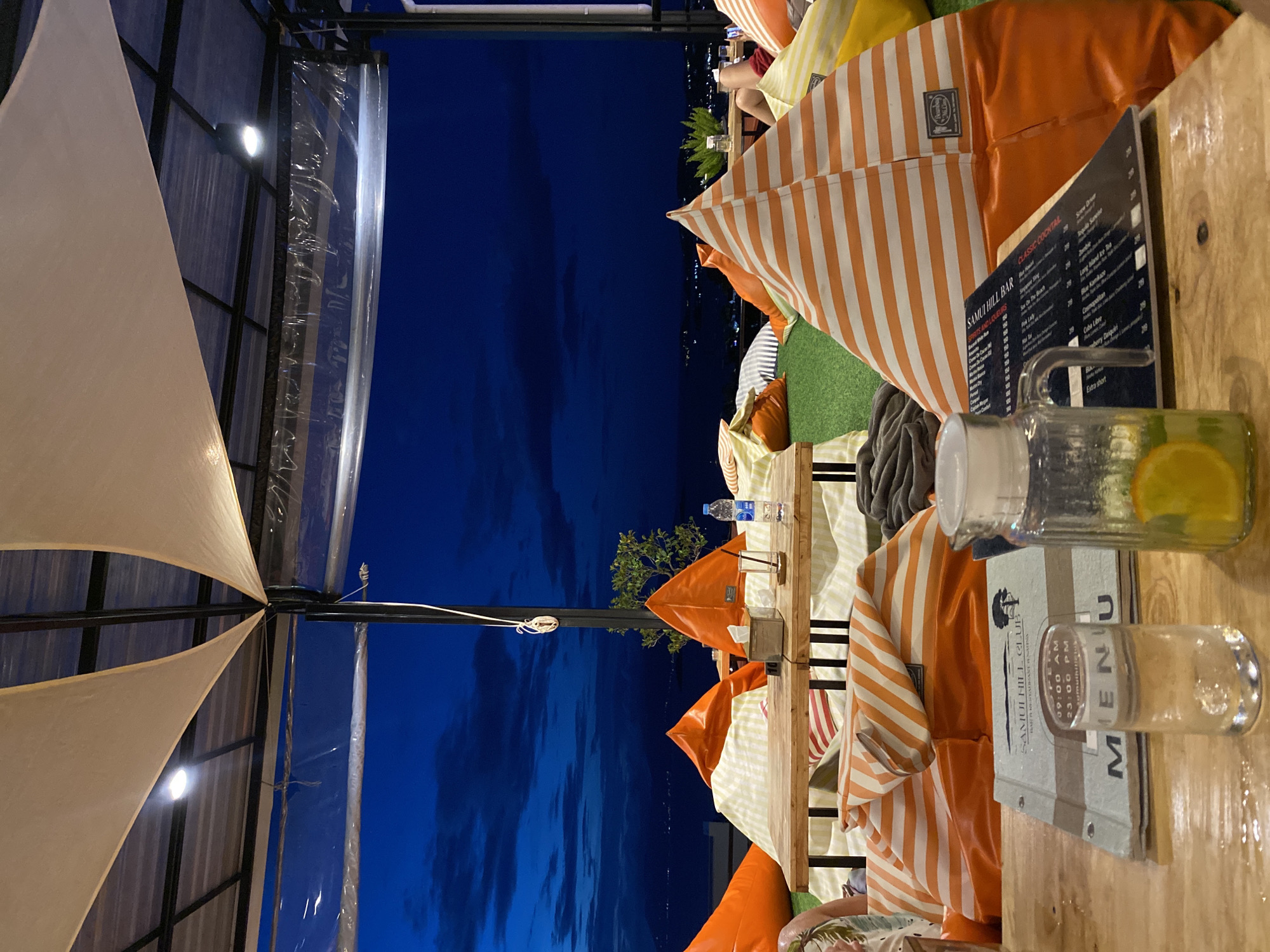 The image captures a scene at Samui Hill Bar in บ้านบางรักษ์, Thailand, at night. Foreground elements include a wooden table, menu, a glass with a lid, and a glass with water and lemon slices, positioned near several orange and white striped beanbag chairs. The environment features a covered outdoor seating area with a view of the night sky. The menu displays the bar's name, operating hours (09:00 AM to 23:00 PM), and a list of classic cocktails along with other items. In the background, a clear plastic wall separates the seating area from the dark outdoor space, where lights suggest the presence of the cityscape. There is a bottle of water sitting on a wooden table behind the table closest to the camera. The scene appears to be an outdoor setup, likely for patrons to enjoy drinks and the view.