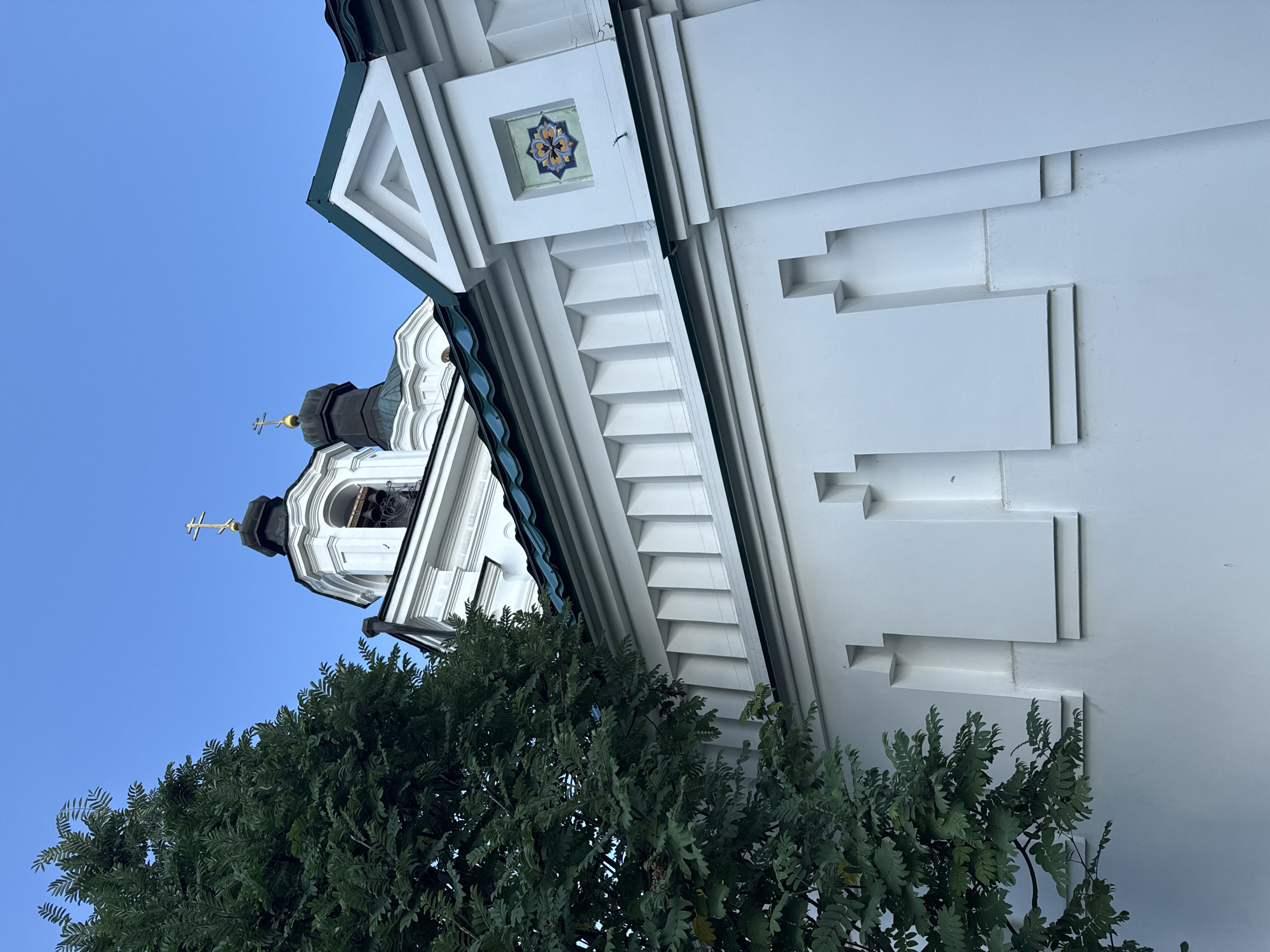 A low-angle shot captures a portion of the white walls and green-tiled roof of an Orthodox church against a clear blue sky. A large tree with dark green foliage dominates the left side of the frame, partially obscuring the church's facade. The church features ornate architectural details, including tiered white walls with decorative elements, tiered roofs, and two dark domes topped with golden crosses. A small, colorful mosaic adorns one section of the white wall. The scene appears to be an exterior view of the Church of the Holy Spirit in Yekaterinburg, Russia.