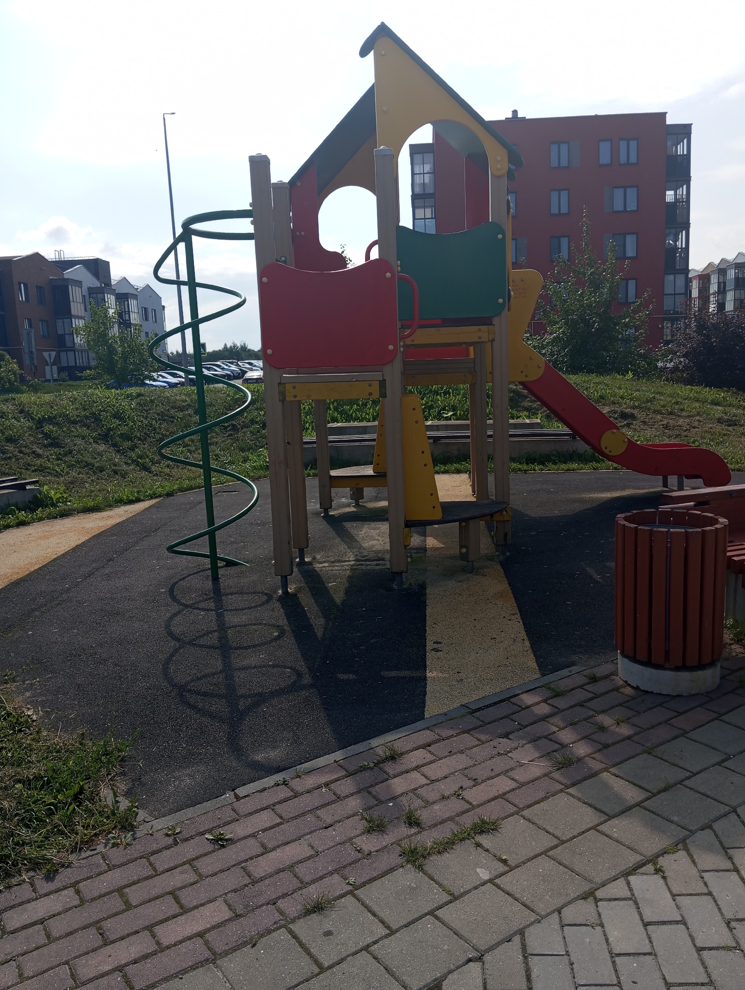 An outdoor playground is depicted with a multi-colored play structure featuring a red slide, a yellow and green roof, and red and green panels. A green spiral slide is attached to the side of the structure. The playground surface is made of dark asphalt with a yellow stripe and is bordered by a brick pathway. A brown wooden trash receptacle is situated near the edge of the pathway. In the background, modern apartment buildings can be seen, along with parked cars and green grass. The sky is bright and overcast.