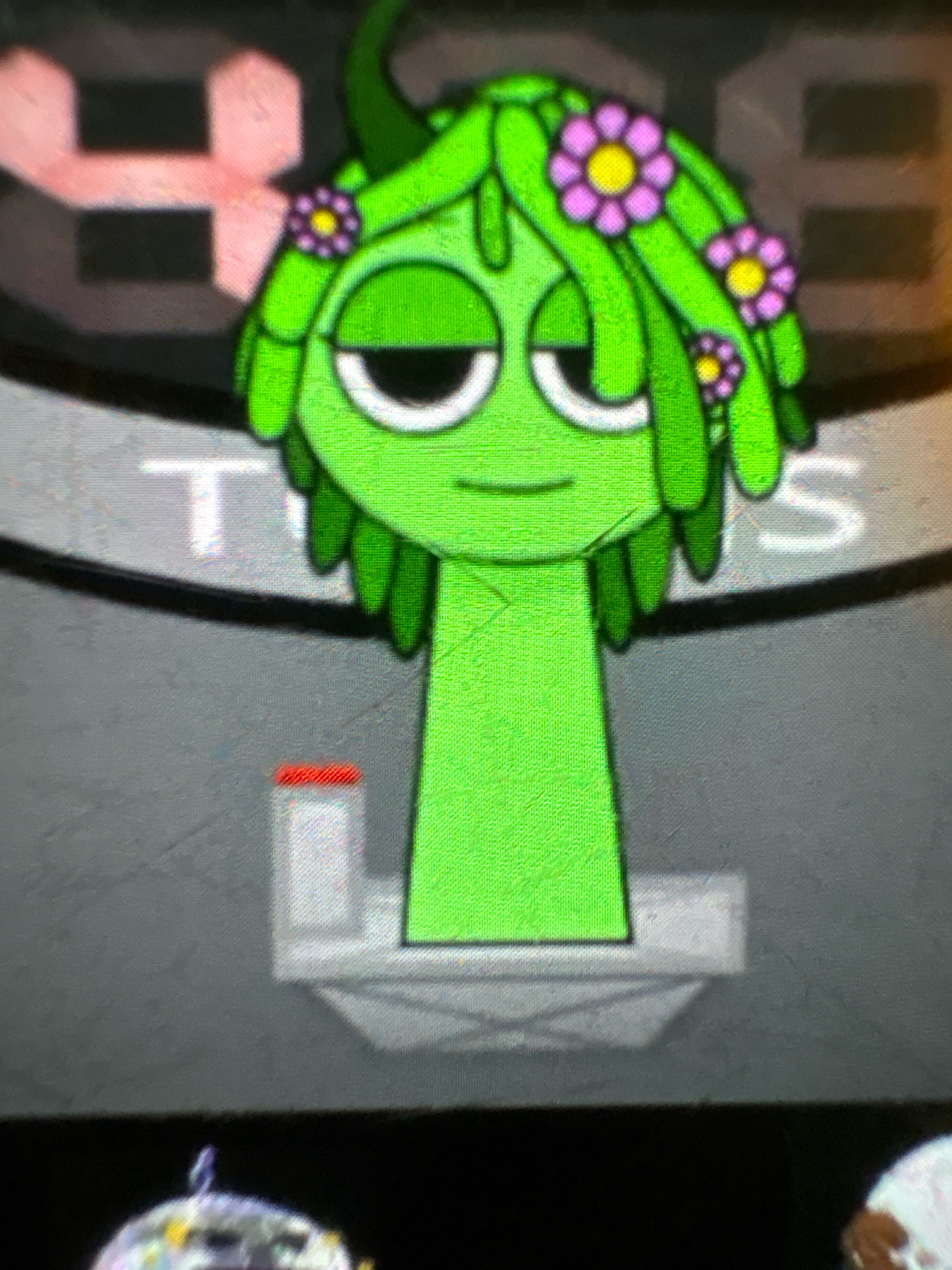 The image displays a cartoonish, green character with long, flowing green hair adorned with small purple and yellow flowers. The character has large, sleepy-looking eyes with white outlines and black pupils. It has a long, rectangular green neck and appears to be standing on a grey, tiered platform. In the background, large, blurry, pink and white shapes are visible, possibly text or graphics. The overall aesthetic is digital and animated. The scene is not located in Ashvem, India.