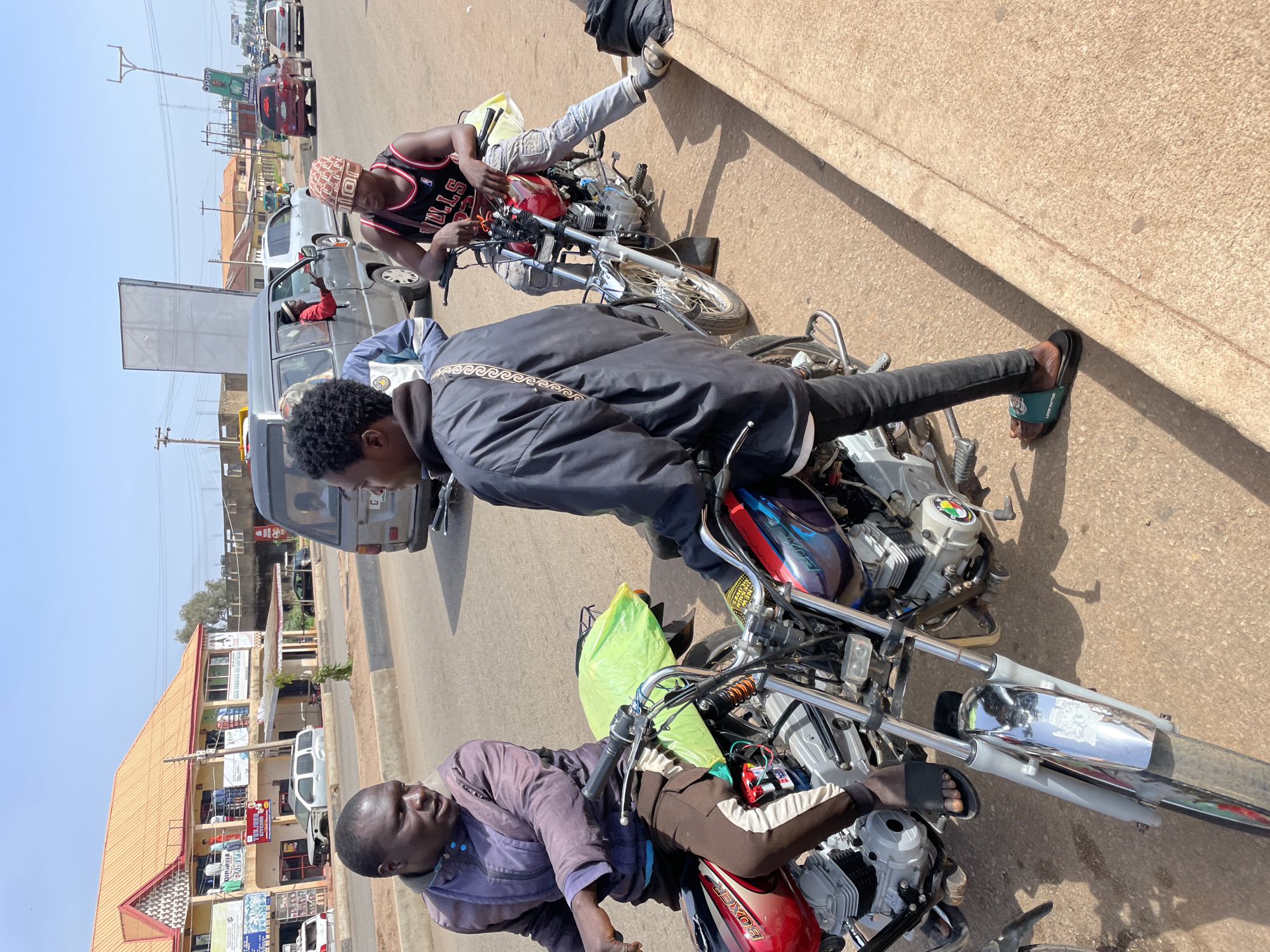The image captures a bustling street scene in Gura Top, Nigeria, under bright midday sun. Three men and several motorcycles dominate the foreground and midground, with buildings and traffic lining a paved road in the background.
In the immediate foreground, a man with a bald head wears a purple long-sleeved shirt and brown trousers, sitting astride a red "BOXER" brand motorcycle. A neon green bag is secured to the back of another motorcycle near him. Slightly to the right and standing, a man with short, curly hair wears a black jacket with a patterned strap, black pants, and green-accented "LOUIS VUITTON" slides, looking downwards. Further right, another man wears a black basketball jersey with "BULLS 23" printed on it and a patterned cap. He sits on a red motorcycle, seemingly adjusting something near the handlebars, dressed in light jeans and sneakers.
The men appear to be pausing or waiting by the roadside, possibly in traffic or a designated stop. Strong sunlight casts distinct shadows, indicating clear weather.
In the midground, a grey van is visible, with a person in a red shirt partially seen inside. Other vehicles are queued further down the street, suggesting a busy road or traffic congestion. Utility poles and overhead wires crisscross the scene.
The background features several multi-story buildings with reddish-brown tiled roofs and light-colored facades. Many commercial signs are affixed to these buildings, some readable including "DUKUMA FAMILY," "AL-JABBAR TRADING CO. LTD," "ZARUMAI & SONS STORE," and "SAFIYA GENERAL STORE." A tall billboard structure displaying the "O'Pay" logo is also visible in the distance. The overall scene depicts a typical vibrant and active urban or semi-urban street environment.