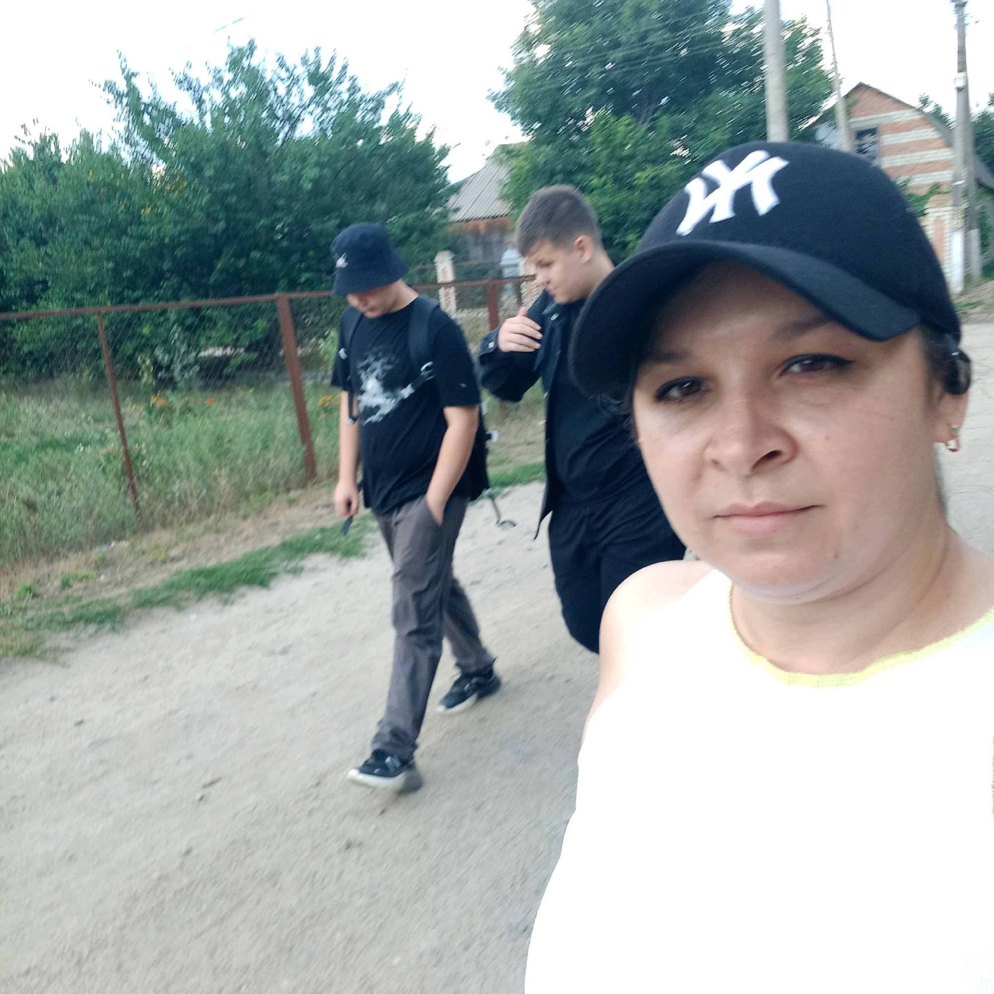 A woman wearing a white tank top and a black baseball cap with a white "NY" logo is in the foreground of the image, looking forward. Behind her, two young men are walking away from the camera on a dirt path. The man on the left wears a dark bucket hat, a black t-shirt with a graphic design, and grey pants. The man on the right wears a black jacket and black pants. A rusty metal fence runs along the left side of the path, with green foliage and trees behind it. In the background, a brick building with a grey roof is visible. The sky is overcast. The scene appears to be in an outdoor, possibly rural, setting.