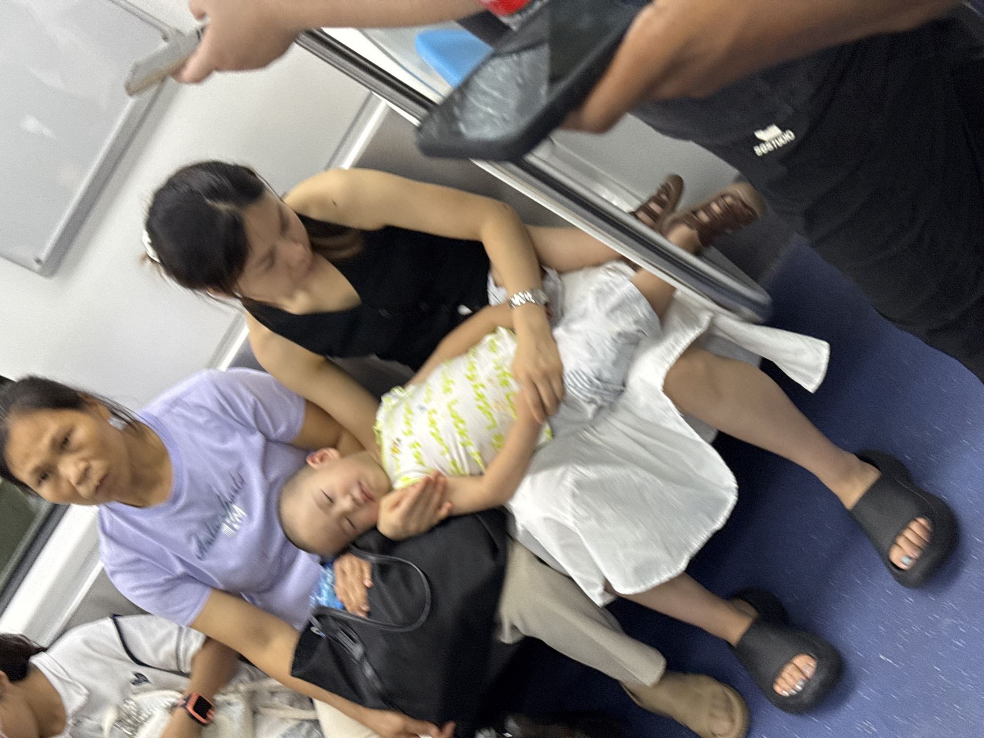 A young boy sleeps soundly in his mother's lap on a public train. He is wearing a white and green shirt and white shorts. His mother, a woman with dark hair pulled back, wears a black sleeveless top. Beside them, another woman, wearing a light purple t-shirt and light-colored pants, sits with a black tote bag on her lap. A third woman, partially visible on the left, wears a white polo shirt. The train interior is visible, with white walls and a blue floor. The lighting suggests it is daytime.