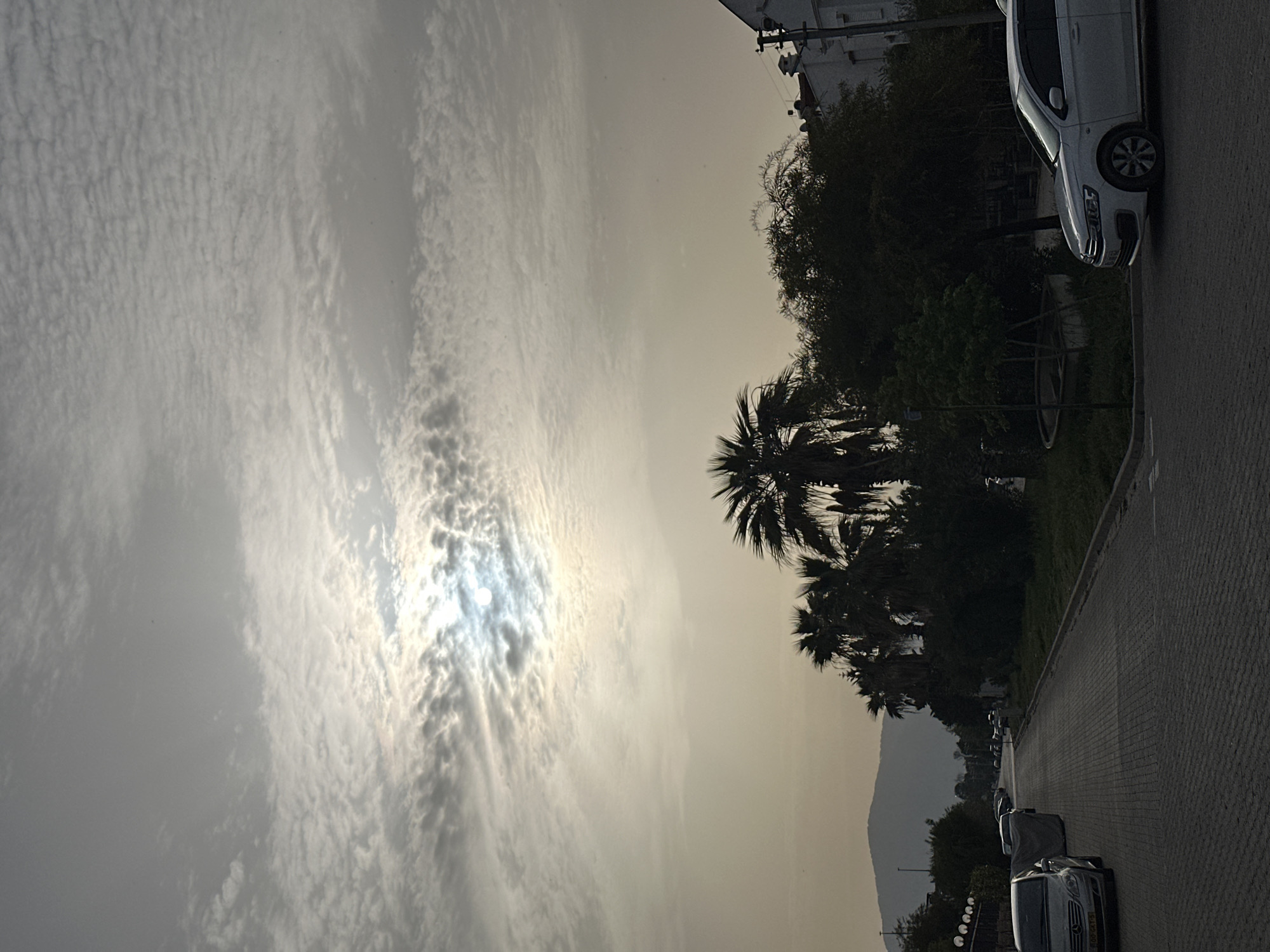 The image depicts a street scene in Fethiye, Turkey, under a sky filled with textured clouds obscuring the sun. Palm trees are visible, their silhouettes contrasting against the sky. Parked cars line the side of the street, with a white van prominently parked near a building. A paved walkway runs alongside the street. In the distance, the silhouette of a mountain is visible.