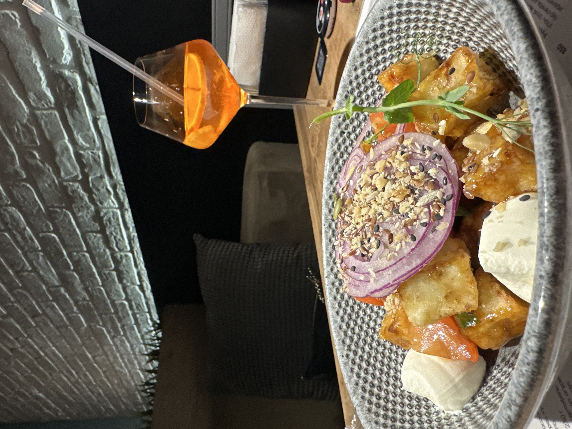 A high-angle shot showcases a plate of food and a drink. The food appears to be a salad with fried tofu, red onion, tomatoes, and a creamy white cheese, possibly burrata or mozzarella, dressed with herbs and seeds. It's served in a textured, patterned bowl. Beside the plate, there's a glass of orange liquid with an orange slice and a clear straw, likely a cocktail. The setting suggests a restaurant or dining area, possibly in the city of Chaikovsky, Russia. Behind the plate and drink, there are hints of a table surface, a neutral-colored chair or cushion, and a dimly lit area. In the far left, a brick-patterned wall is visible. The overall impression is that of a contemporary dining experience.