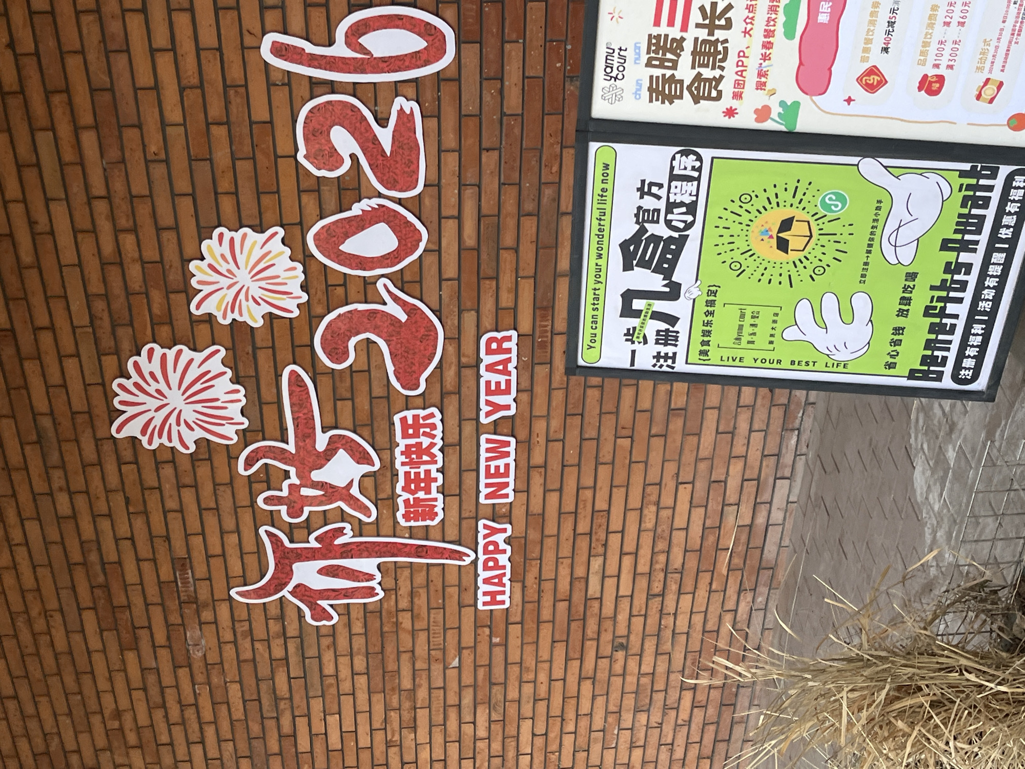 This image shows a wall advertising Chinese New Year (2026) and a "Yamu Court" food-related app and mini-program in Changchun City, China. The wall is made of brick, with the numbers "2026" and the characters for "Happy New Year" affixed to it in a stylized, red-and-white design. Below this is the English phrase, "HAPPY NEW YEAR" in block letters. There are also firework designs on the wall.
To the right of the New Year decorations is a framed advertisement for a "Yamu Court" mini-program for food and entertainment. The poster mentions "Chun Nuan San Shi Hui Chang," indicating a Spring warmth catering fair. It also mentions searching for the app on Meituan and Dazhong Dianping. The ad features cartoon hands and encourages users to "live your best life" by registering for the app. Offers and discounts are listed in yuan (currency) for the app.
The scene is outside and includes some dry vegetation near the bottom of the frame. The overall impression is a promotion for both the upcoming New Year and a local food app.