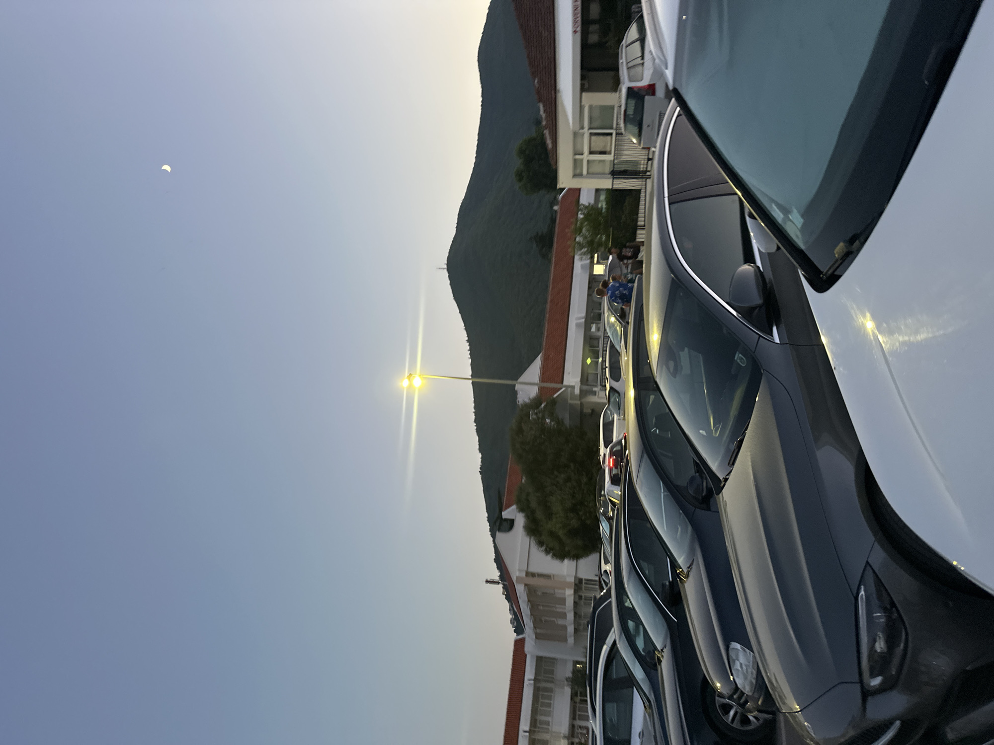 In Budva, Montenegro, a parking lot is depicted at dusk, with a crescent moon visible in the clear, pale blue sky. Several cars are parked in rows. In the background, a building with red-tiled roofs and white walls is visible, situated at the base of a lush, green, tree-covered mountain. A street lamp with bright lights illuminates the scene. A few people, including two young males and one adult male, are standing near the building on the left side of the image.