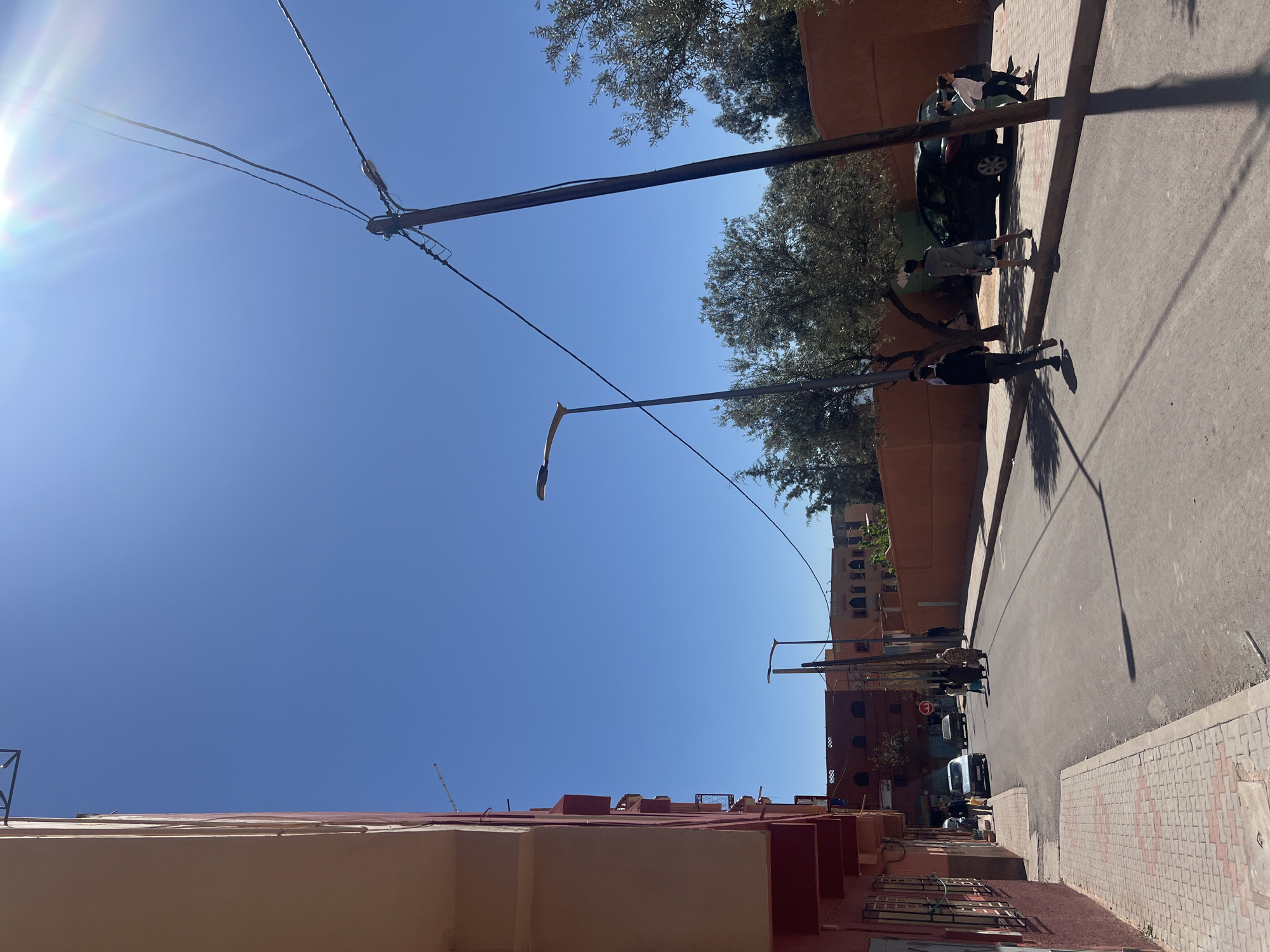 The image shows a street scene in Ouaouizert, Morocco on a sunny day. Buildings with red and peach-colored exteriors are visible on the left side of the frame. The street is paved and partially shaded. Three people are walking along the sidewalk on the right side of the street. Several cars are parked along the street, including a dark-colored car and a white van. A red brick wall runs along the side of the street. Utility poles and wires span across the sky. Trees are interspersed among the buildings.