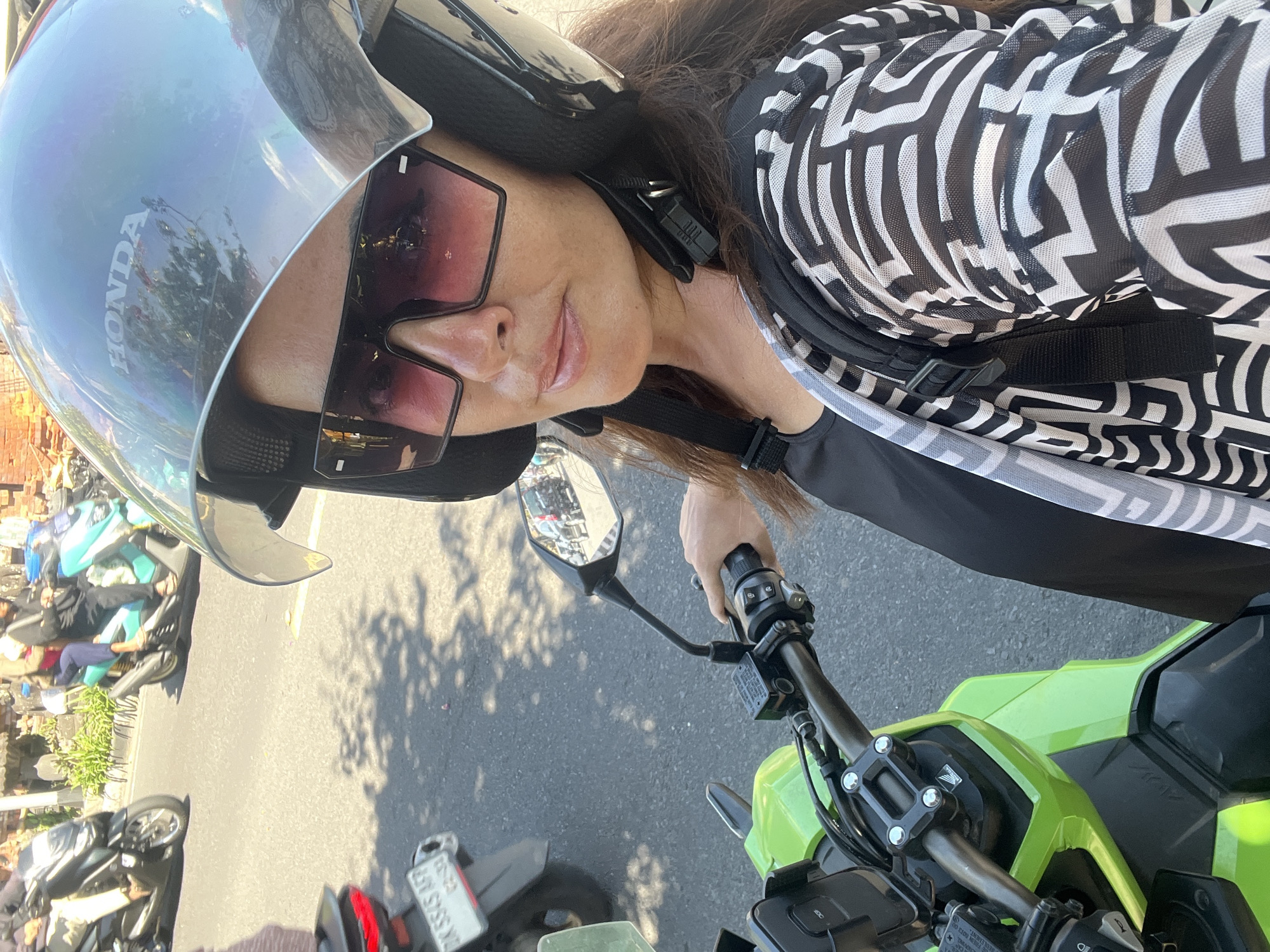 A woman wearing a helmet and sunglasses is riding a bright green scooter down a street in Sukawati, Indonesia. She is looking at the camera and smiling. The street is busy with other scooters and motorbikes. There are shops and buildings on either side of the street. The woman is wearing a black top and a black and white patterned jacket. She has a backpack on. The sun is shining and the sky is blue.