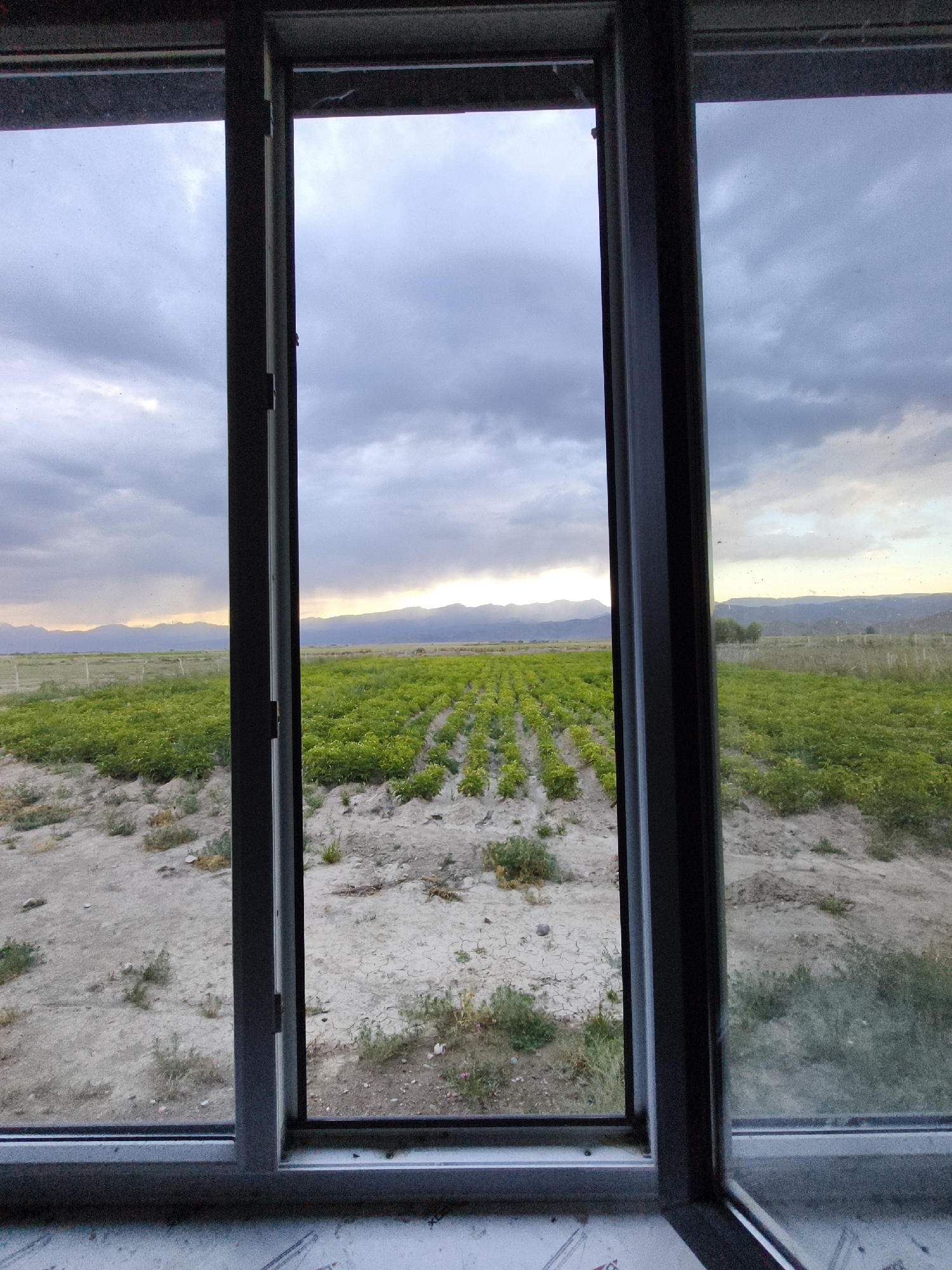 A low-angle, eye-level shot captures a view of a field through an open window. The window frame, dark gray, bisects the scene vertically, with the left pane slightly ajar. Outside, a vast expanse of green crops stretches towards distant mountains under a cloudy sky. The foreground consists of dry, cracked earth with sparse vegetation. The sky is dramatic, with dark clouds dominating the upper half, but shafts of sunlight break through, illuminating the mountain range in the distance with a warm glow. The overall impression is one of a rural landscape observed from indoors.