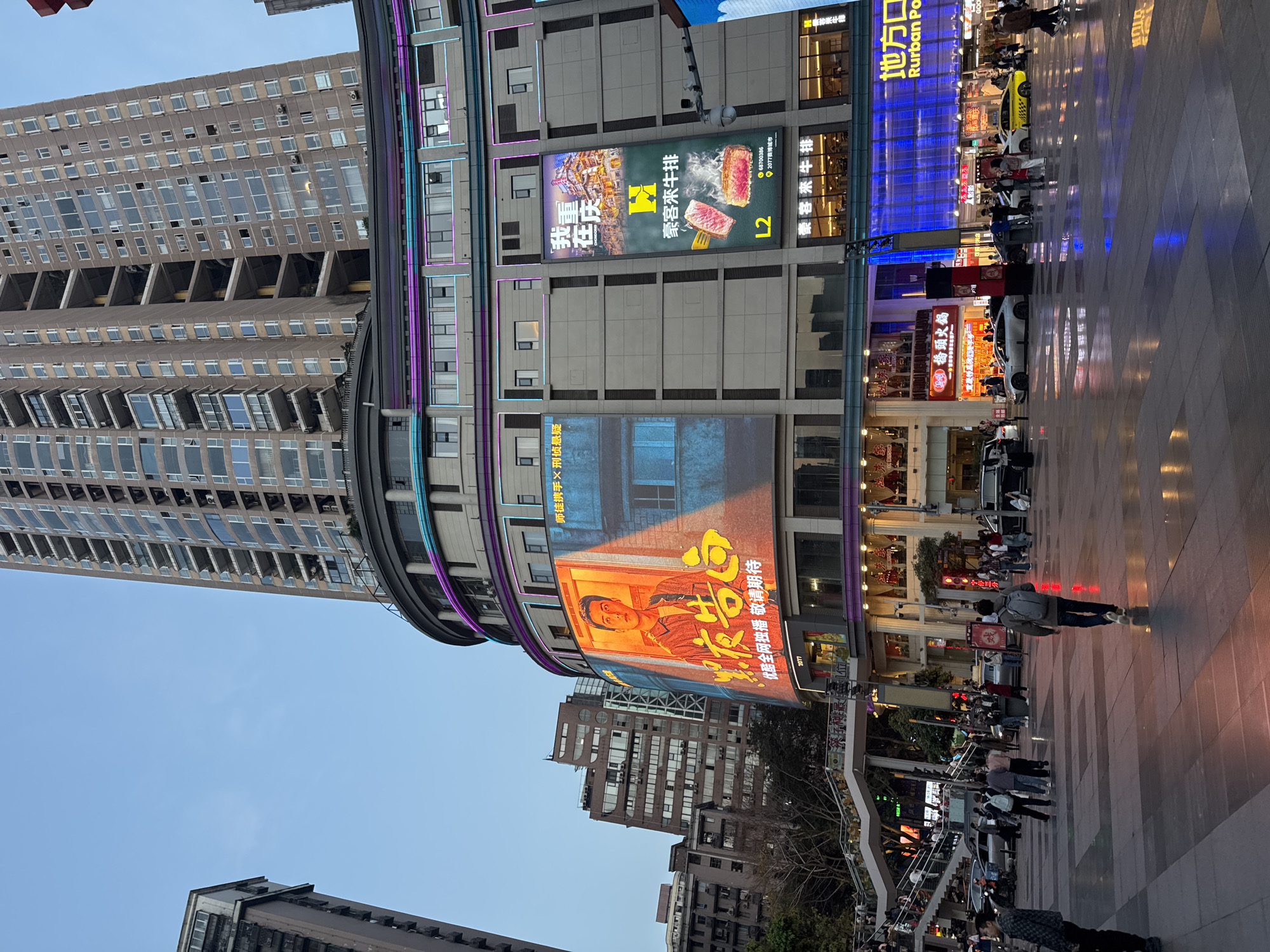 The photograph captures a busy urban scene in what appears to be the Yuzhong District of Chongqing, China. The sky is a clear blue. A tall, modern building with a curved facade is prominently featured, adorned with large LED screens displaying advertisements and entertainment.
A large ad features a male figure in an orange-toned graphic, alongside Chinese characters. Other ads include a food promotion for "豪客来牛排," which translates to "House of Steak", and a shop named "Rurban Port."
Below the building, a pedestrianized area is bustling with people. The ground is paved and reflects the bright lights of the surrounding establishments. A white taxi and other vehicles are interspersed among the pedestrians. There's a complex arrangement of stairs and architectural structures in the middle ground, with more buildings visible in the background, partially obscured by the closer structures. The overall impression is one of a lively commercial district.