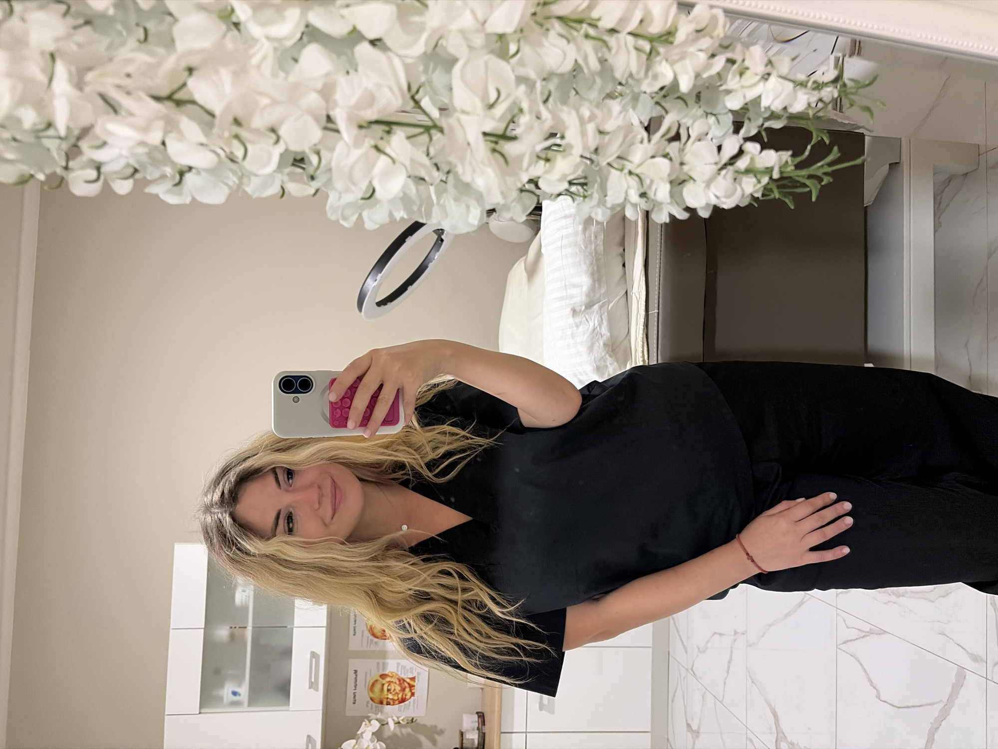 A woman with blonde hair, wearing black scrubs, stands in a room, taking a selfie with her phone. She is smiling slightly and looking at her phone. The room appears to be a medical or aesthetic clinic. In the background, there are white cabinets with medical supplies, a treatment bed, and a ring light. A large white floral arrangement hangs in the foreground, partially obscuring the view of a mirror. The floor is tiled with a marble pattern. The location is identified as Budva, Montenegro.