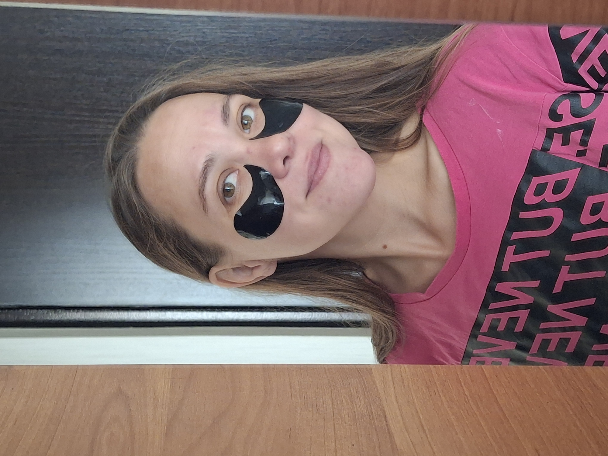 A young woman with light brown hair is pictured from the chest up, positioned on the right side of the frame. She is wearing a bright pink t-shirt with large black lettering that reads "NEVER BUT NEVER". Black under-eye patches are affixed to her face. Her skin appears fair with some light freckling. She is looking towards the camera with a slight smile. The background consists of a doorway, with a wooden door on the left and a darker door on the right, suggesting an indoor setting in Chaikovsky, Russia. The lighting appears to be natural, coming from the left.