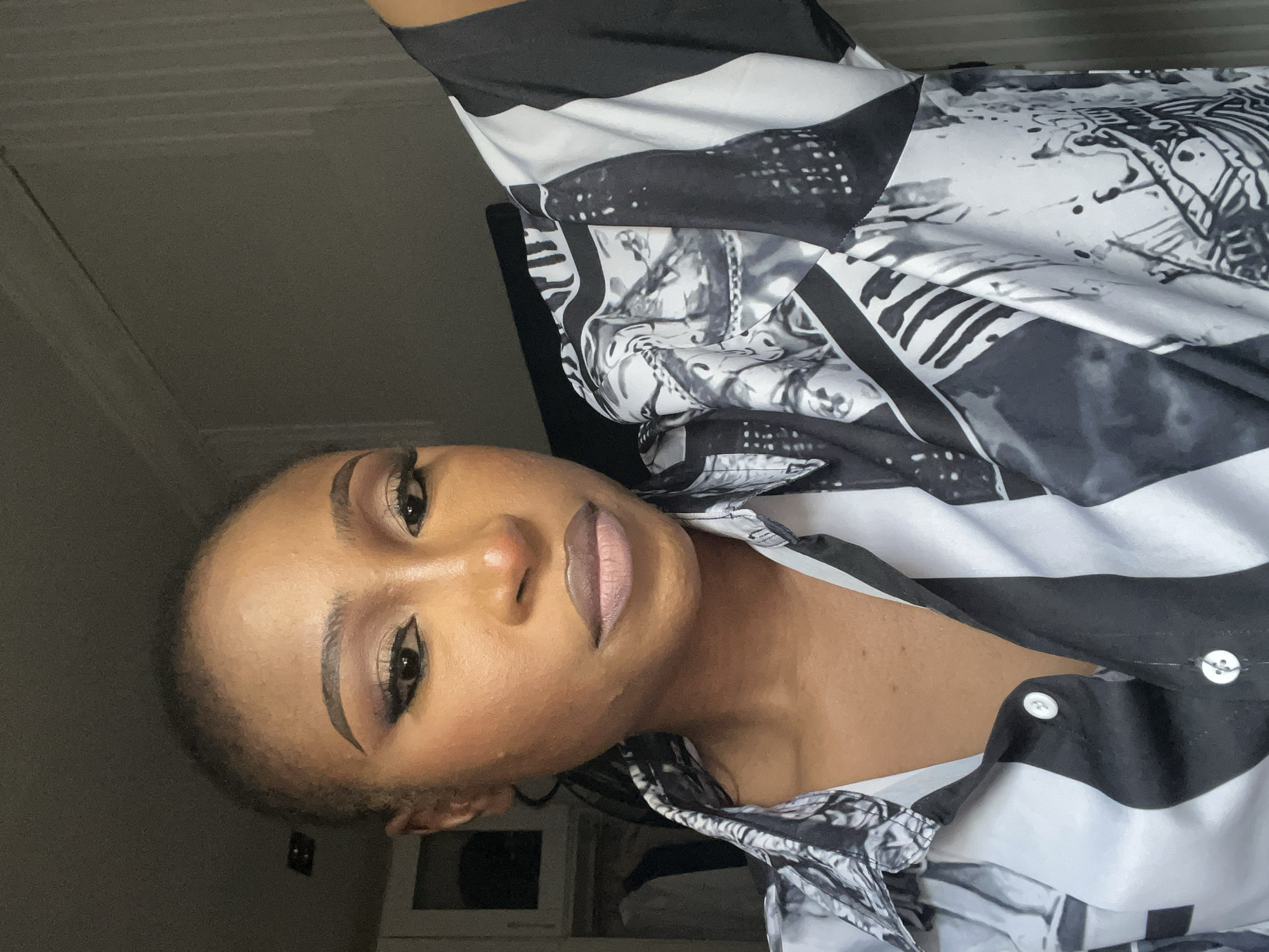 This is a close-up, selfie-style portrait of a Black woman indoors, likely in a residential setting in Abuja, Nigeria. She has her hair pulled back and is wearing a black and white patterned button-up shirt. Her makeup is prominent, featuring winged eyeliner, eyeshadow, and a nude-toned lipstick. The background shows a wall and part of a wardrobe or closet. The lighting appears to be natural, coming from a source to her left, casting subtle shadows.
