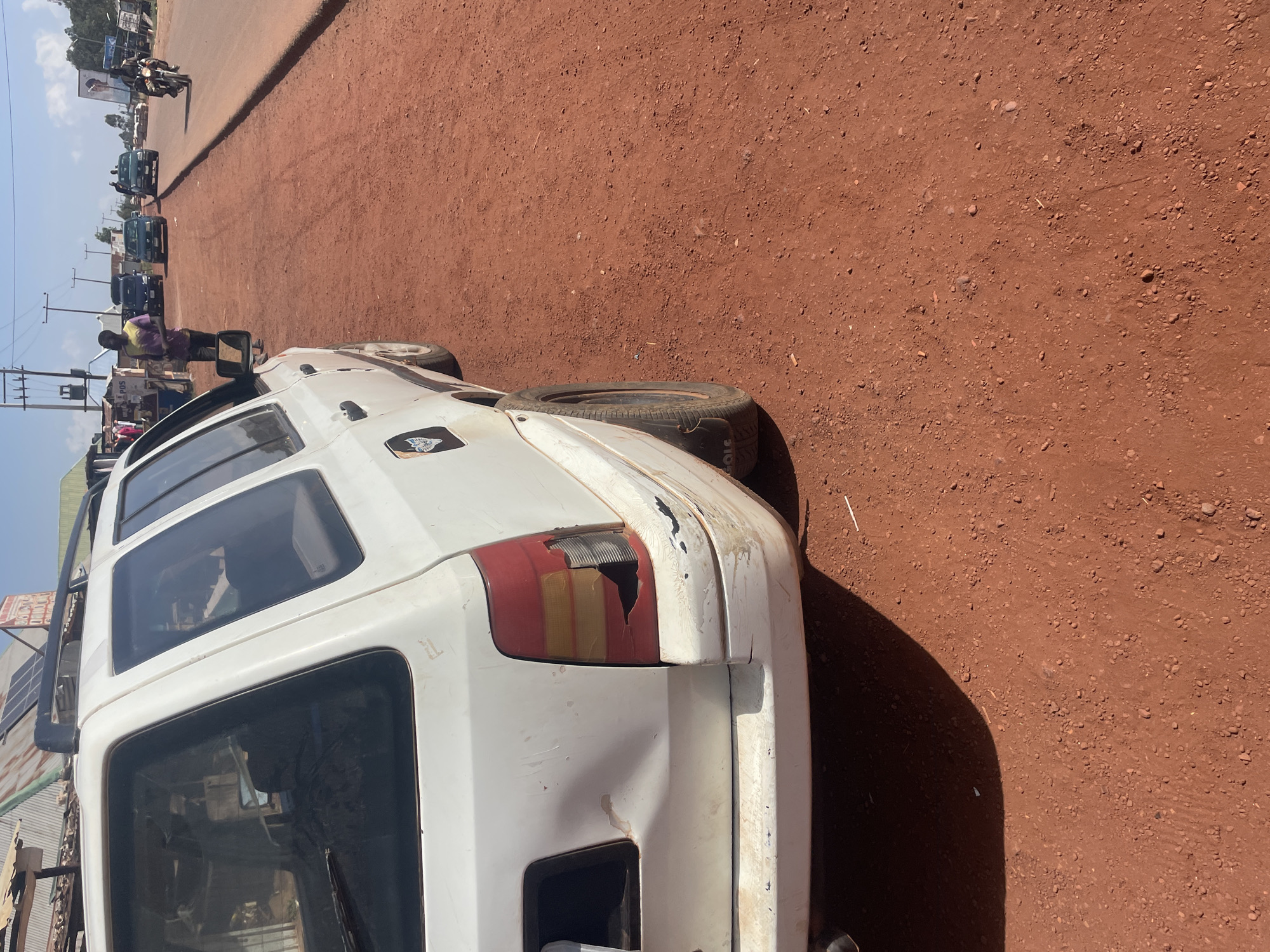 The image captures a roadside scene in Durumi, Nigeria, under bright daylight. The primary subject is a weathered white van, possibly a Toyota Hiace or similar model, parked or stopped on a red-earth, unpaved road. The van exhibits visible damage on its rear passenger side, including a broken and cracked taillight with exposed internal wiring, and a dented bumper. A spare tire is mounted on the roof rack, and a dark sticker or logo is affixed to the side of the van near the front door.
The road is made of reddish-brown earth mixed with small stones and pebbles, suggesting an unpaved or dirt thoroughfare. Strong shadows cast by the van and other objects indicate a sunny day. In the background, several other vehicles, including cars and at least one motorcycle with a rider, are visible along the road. A man wearing a striped yellow and purple shirt and dark pants stands near the front passenger side of the white van. Utility poles and overhead wires line the side of the road, and various structures, including small shops or stalls, are present in the distance. One sign in the background clearly displays the text "POS". Faintly visible text "2010" can be seen on the sidewall of the spare tire on the van's roof. The scene depicts a typical street environment in a semi-urban or rural Nigerian setting.
