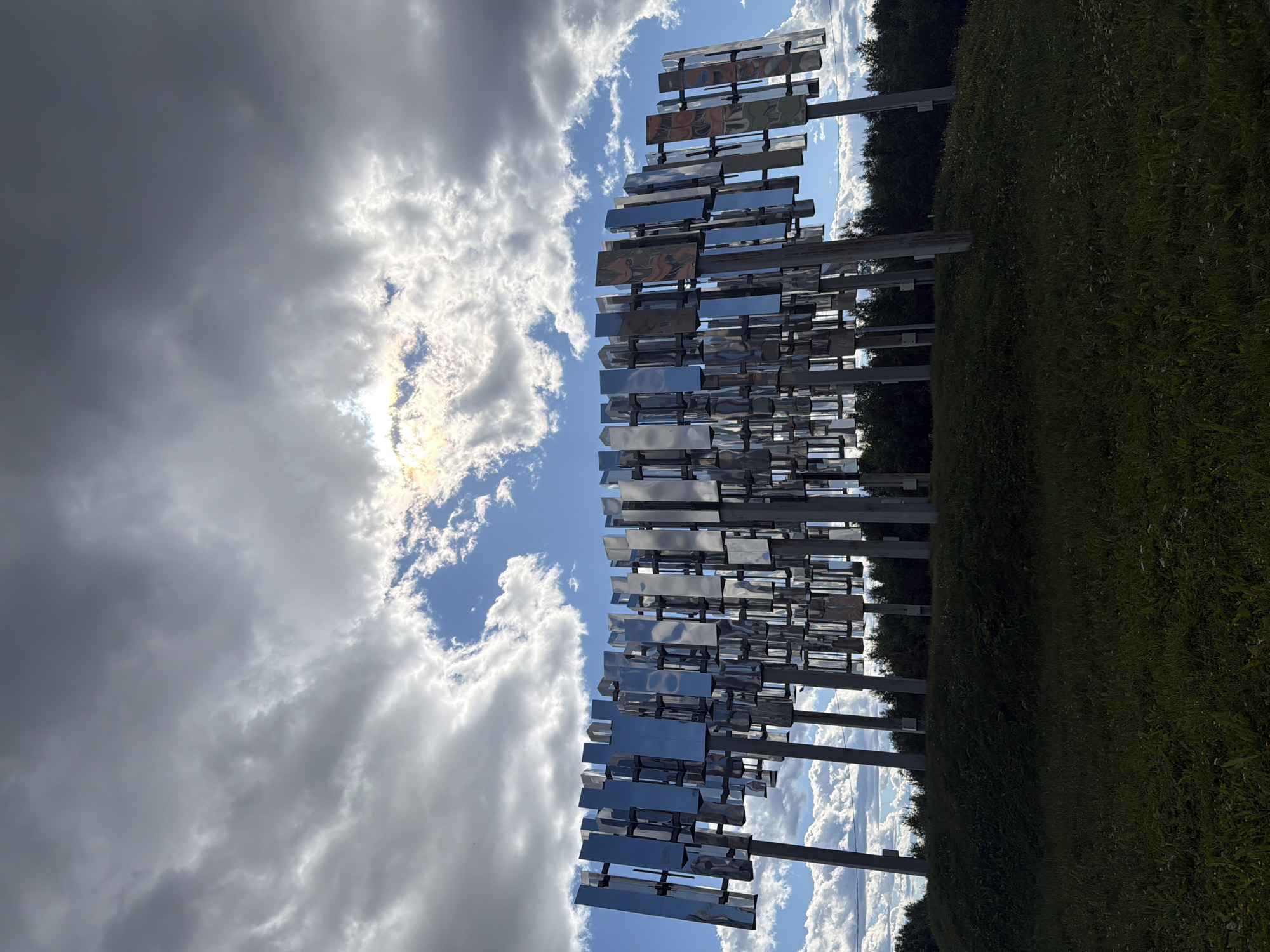 An outdoor, low-angle, full shot shows a large, modern, metallic sculpture positioned on a grassy knoll. The sculpture consists of numerous vertical, rectangular, reflective panels supported by posts, arranged in a curved formation. The panels appear to be made of polished metal, reflecting the sky and clouds. Behind the sculpture, a line of dark green trees forms a backdrop. The sky above is dramatic, with a mix of dark, heavy clouds and bright, sunny patches where the sun is peeking through. This scene is located in the city of Polarnie Zori, Russia. The foreground is dominated by dark green grass, with some small white wildflowers visible. No people or animals are present in the frame.