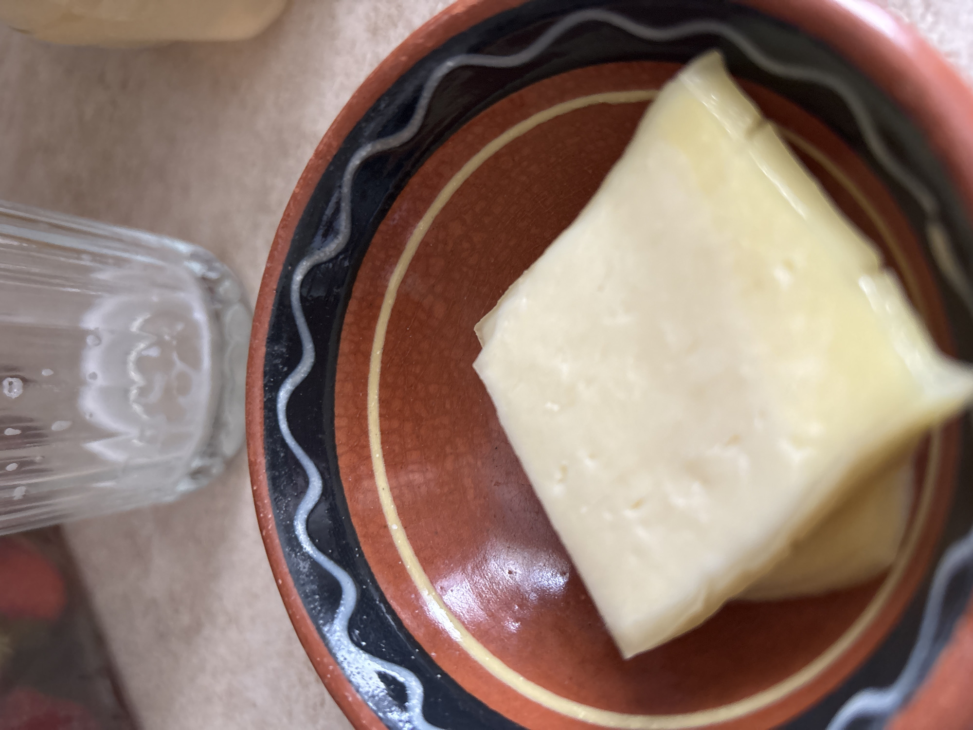 A close-up, overhead shot shows a piece of light yellow cheese, wrapped in translucent plastic, resting in a rustic, earthenware bowl. The bowl has a decorative rim with wavy patterns in black, white, and a reddish-brown color that matches the bowl's interior. The cheese has a smooth, slightly waxy appearance and casts a subtle shadow within the bowl. To the left of the bowl, a portion of a clear, ribbed glass is visible, appearing to be empty or containing a light-colored liquid. The background is a neutral, textured surface, possibly a tablecloth or countertop, suggesting an indoor setting, perhaps a kitchen or dining area. The lighting appears to be natural, casting soft shadows. The image has a shallow depth of field, with the cheese and the bowl being the sharpest elements. No people or animals are visible, and there is no visible text in the image. The overall impression is a simple, still-life composition focused on food preparation or presentation.