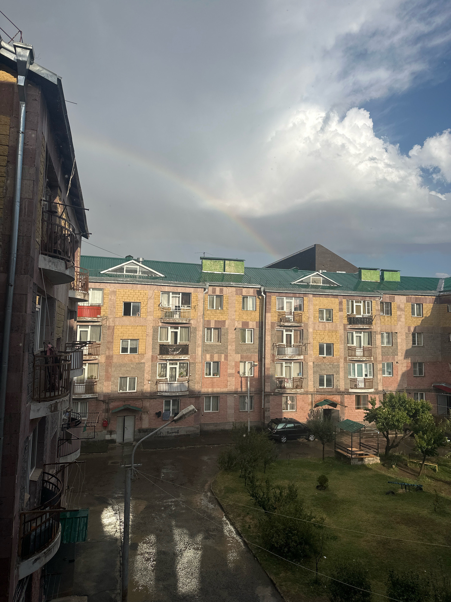 The image depicts an apartment building in Gyumri, Armenia, after a rain shower, with a faint rainbow visible in the sky. The building is made of pink and yellow brick, with green metal roofing and balconies. The ground is wet, reflecting the sunlight and the surrounding buildings. A street lamp stands in the foreground, casting long shadows. There is a small green courtyard with a picnic shelter and trees. A black car is parked in front of the building. The sky is partly cloudy, with a mix of sun and dark clouds, suggesting the rain has just passed. There are no people clearly visible in the scene.