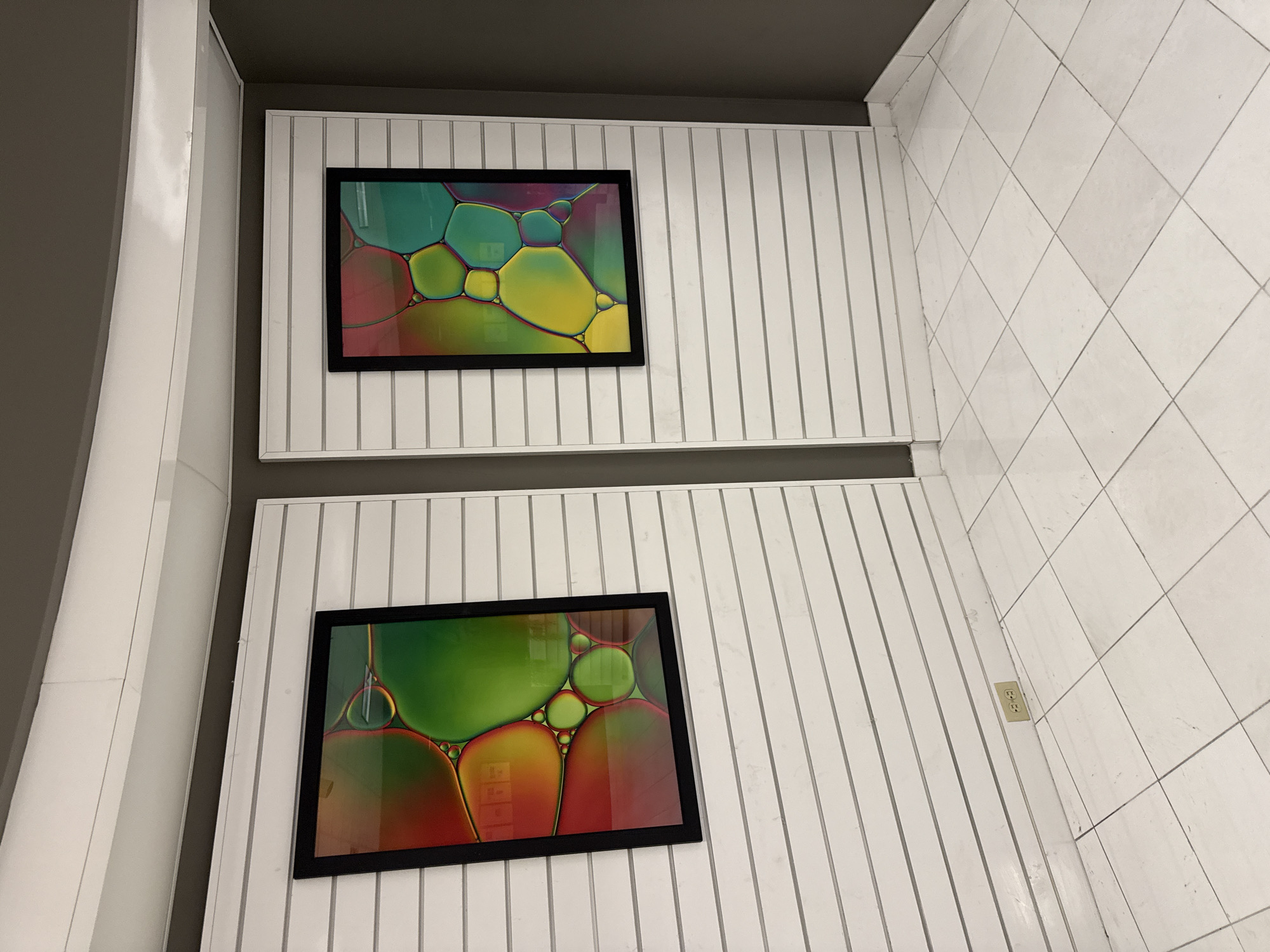 The image shows a close-up of two framed, colorful abstract art pieces hanging on a wall. The wall is constructed of vertical white slats, reminiscent of beadboard paneling. The art pieces, framed in black, feature an abstract design of overlapping circles with a vibrant color palette including greens, yellows, reds, and purples. The ceiling above is dark gray. To the right, the wall transitions to white square tiles. A single electrical outlet is visible on the slatted wall. The perspective suggests the photo was taken at an angle, looking upward. Without additional context, the specific location in Columbus, Ohio cannot be definitively determined.